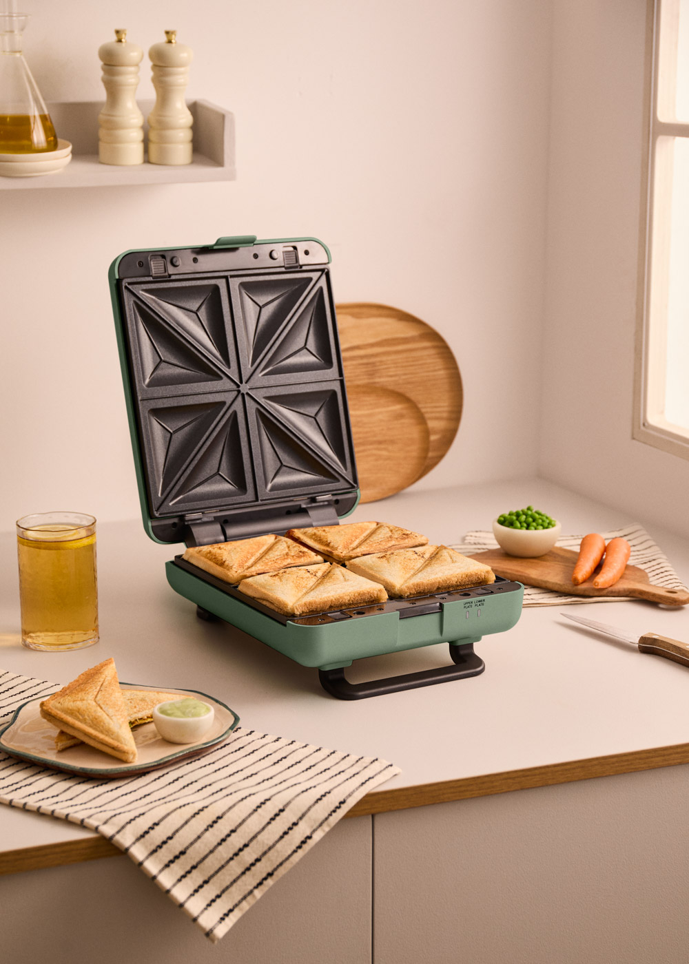 Una sandwichera verde con cuatro sándwiches, un vaso de té y un plato con dos sándwiches cerca.