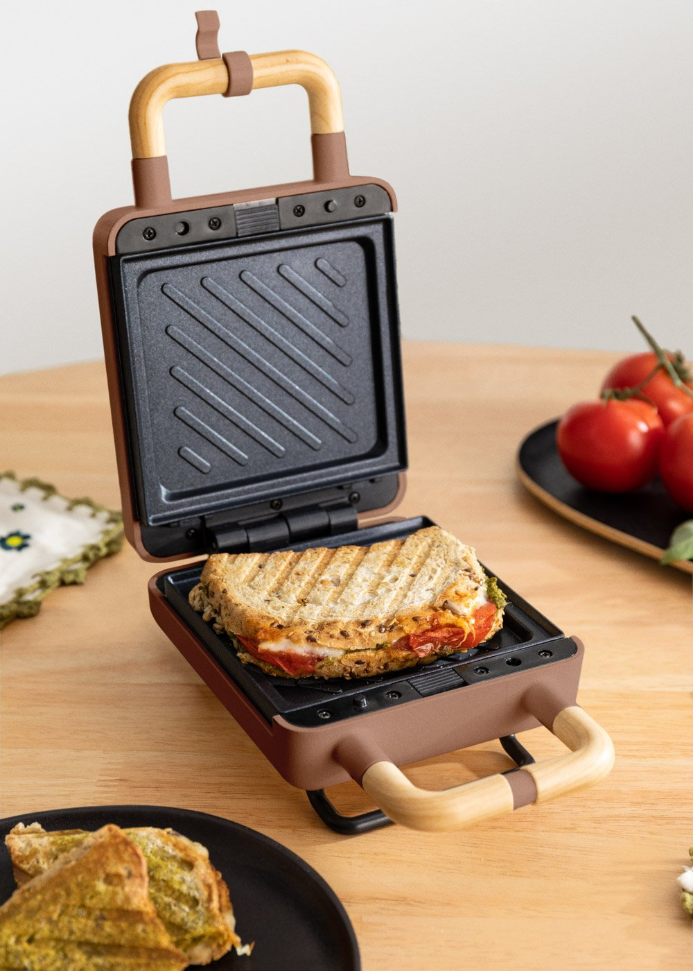 Una sandwichera abierta con un sándwich dentro. Tomates al fondo.