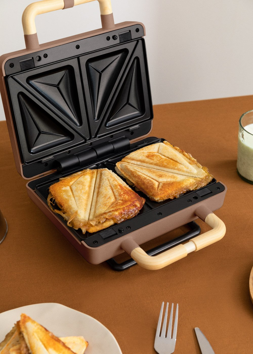 Una sandwichera abierta con dos sándwiches dorados, sobre un mantel marrón.