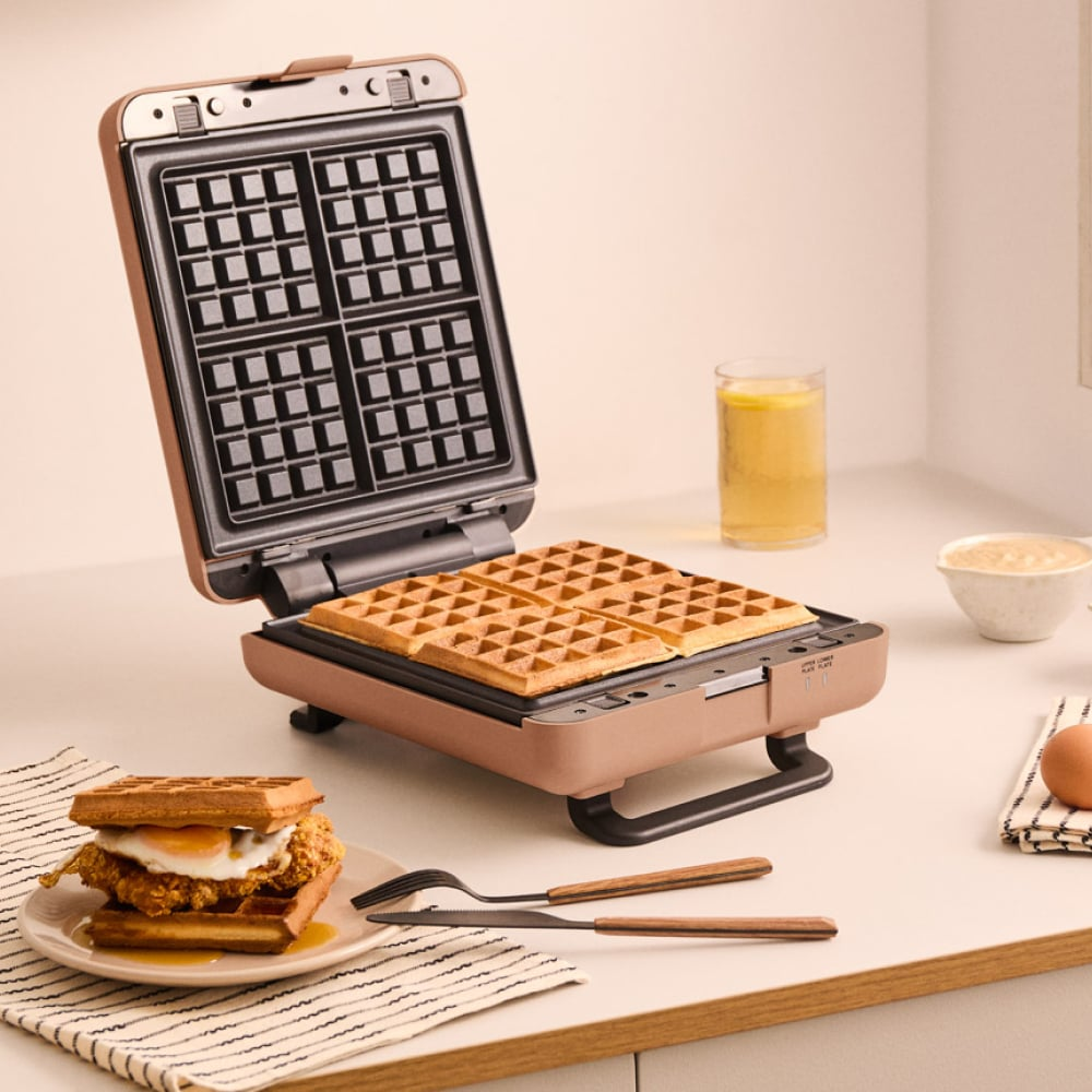 Una gofrera abierta con cuatro gofres cocidos, junto a un sándwich de gofre con huevo y pollo.