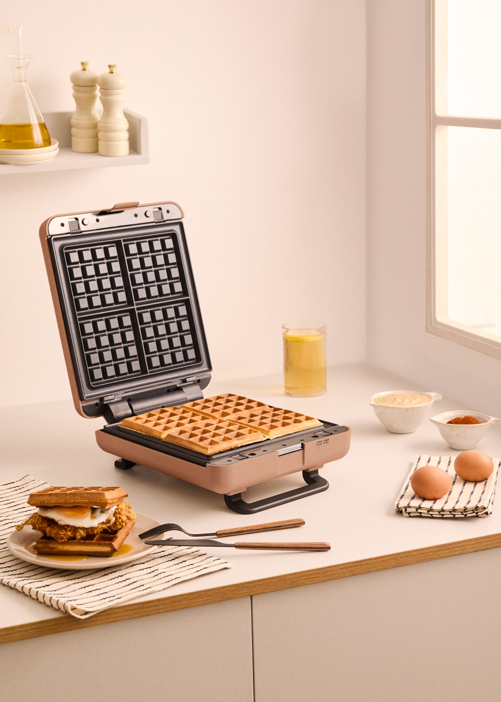 Una gofrera abierta con cuatro gofres cocidos, junto a un sándwich, huevos e ingredientes.