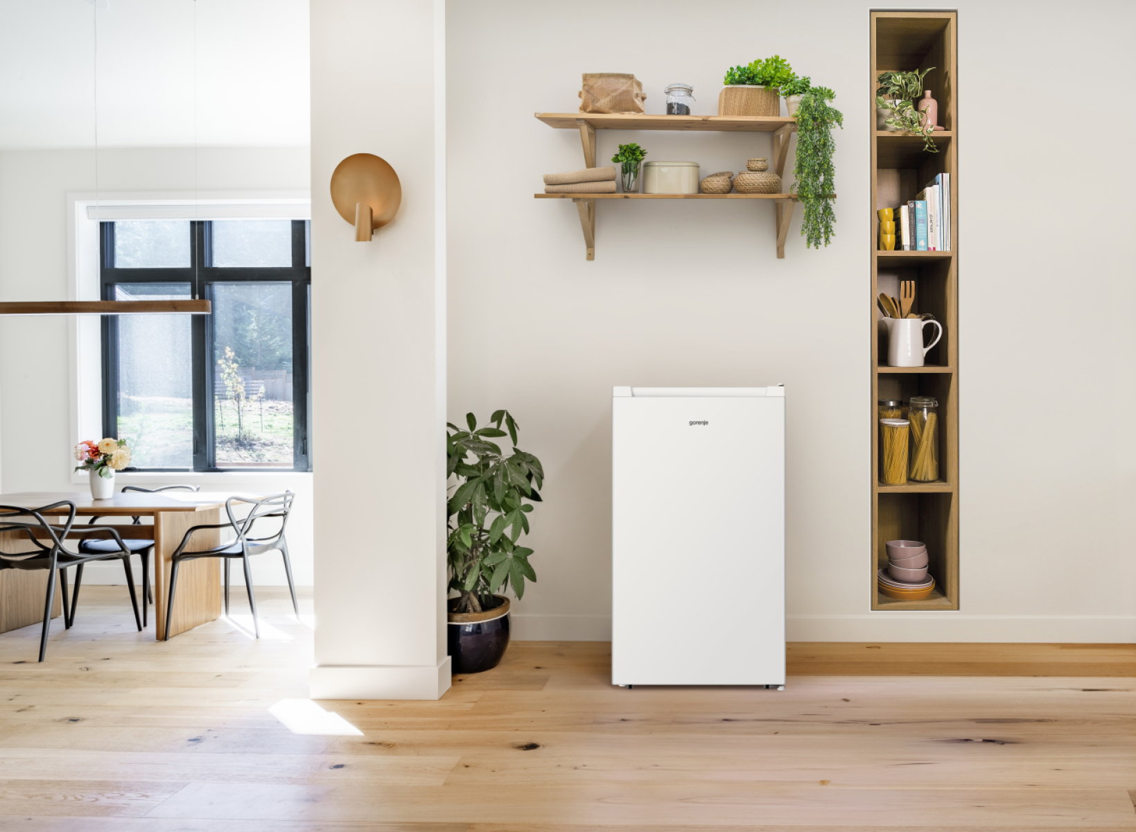 Ein weißer Gorenje-Kühlschrank steht auf hellem Holzboden neben einem hohen Regal.