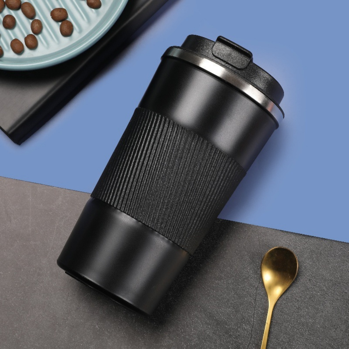 Ein schwarzer Reisebecher mit geripptem Griff steht neben einem goldenen Löffel und Kaffeebohnen.