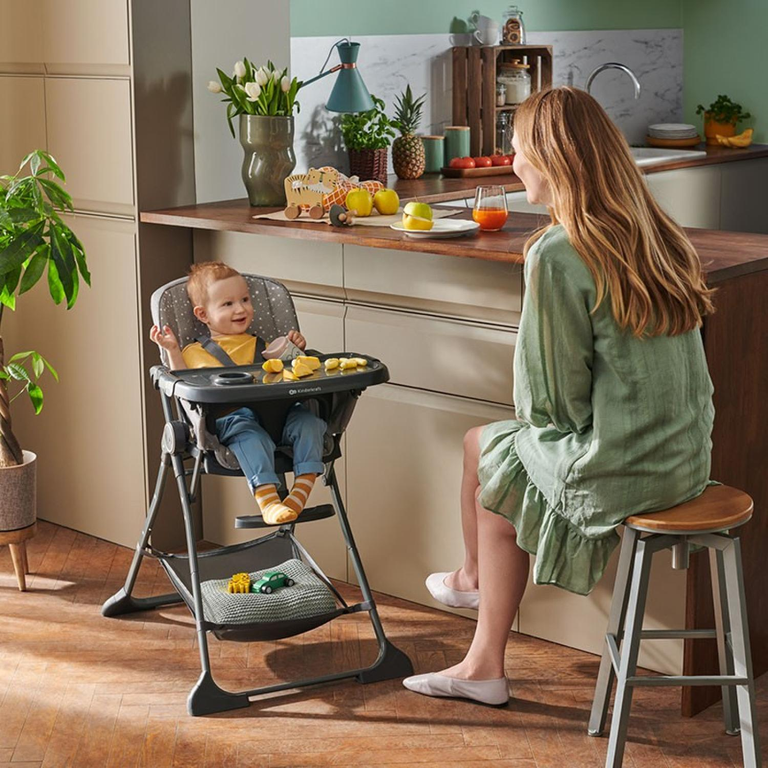 Ein Baby sitzt in einem Hochstuhl, eine Frau sitzt auf einem Hocker daneben.