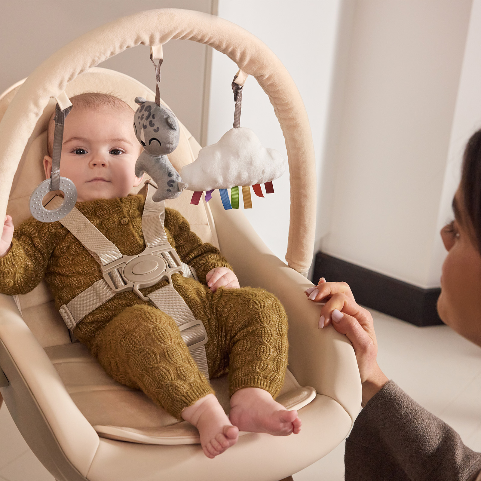 Ein Baby in einem beigen Stuhl mit Spielzeug über sich. Eine Hand berührt den Stuhl.