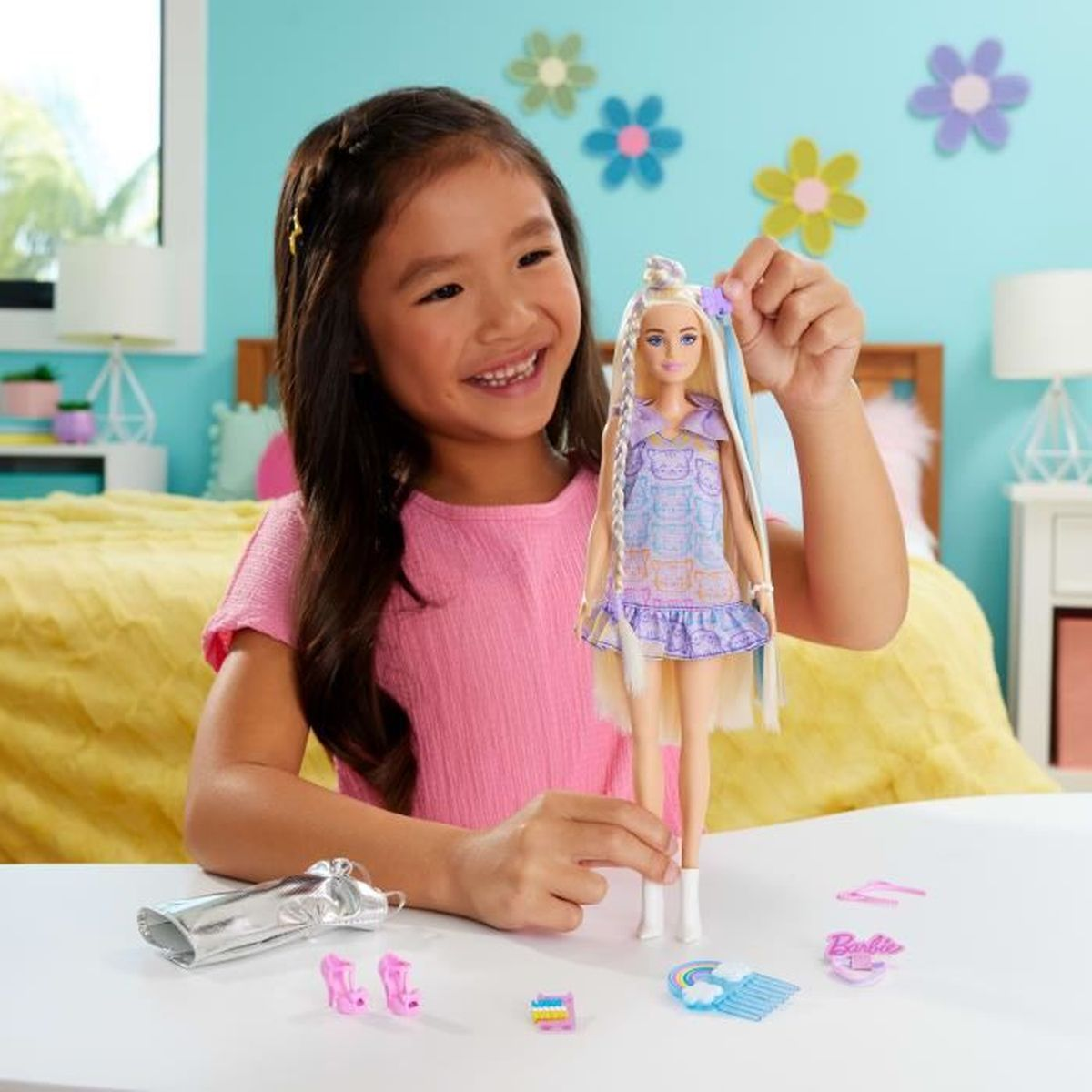 Ein lächelndes Mädchen hält eine Barbie-Puppe in einem blauen Kleid. Zubehör liegt auf dem Tisch.