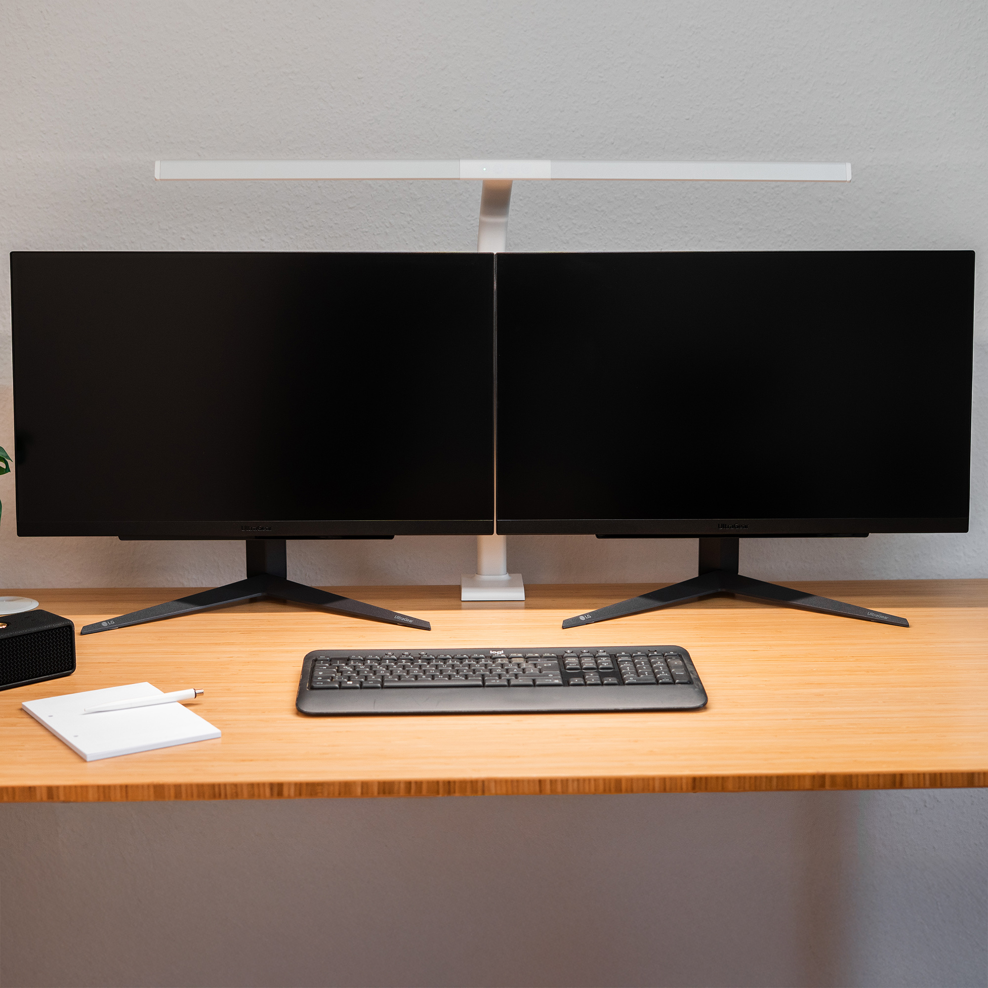 Doppio monitor nero su scrivania in legno con tastiera e blocco note.