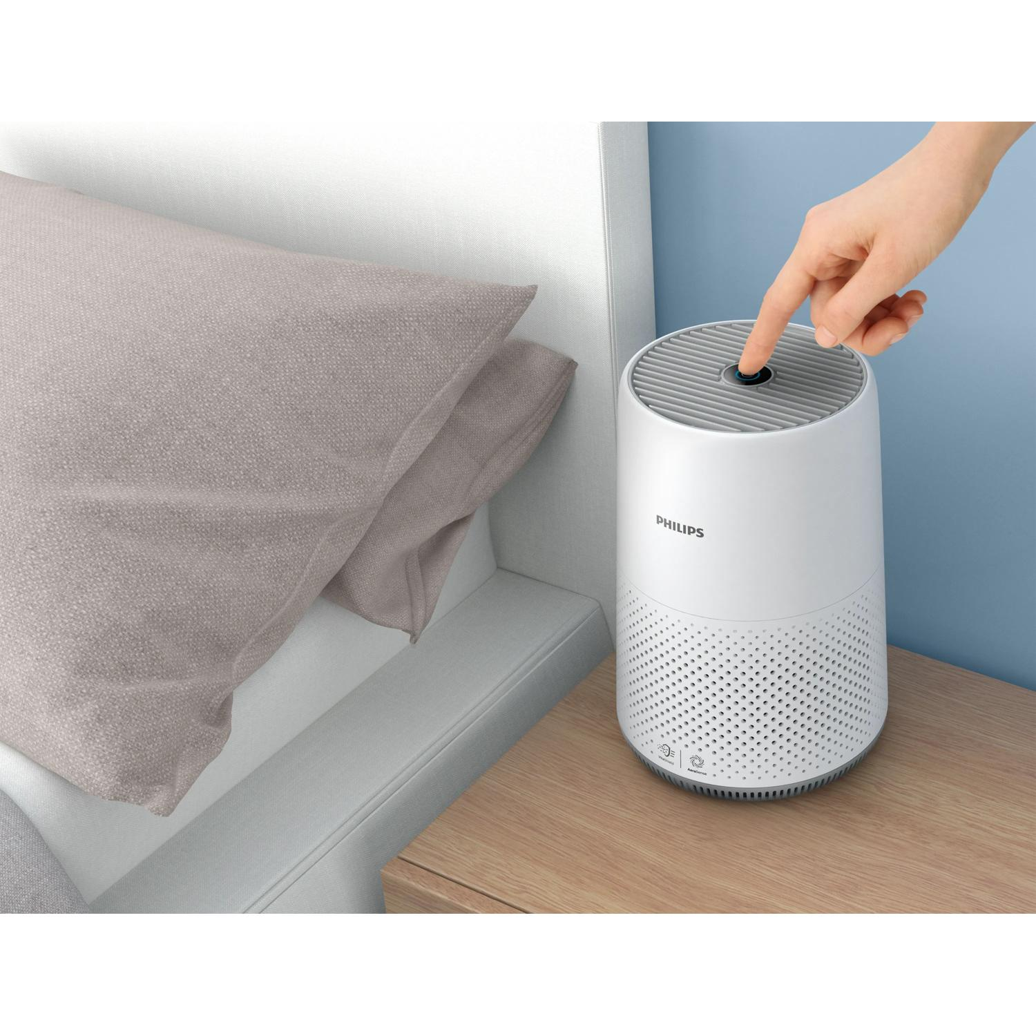 Una mano preme il pulsante di accensione di un purificatore d'aria Philips bianco accanto a un letto.