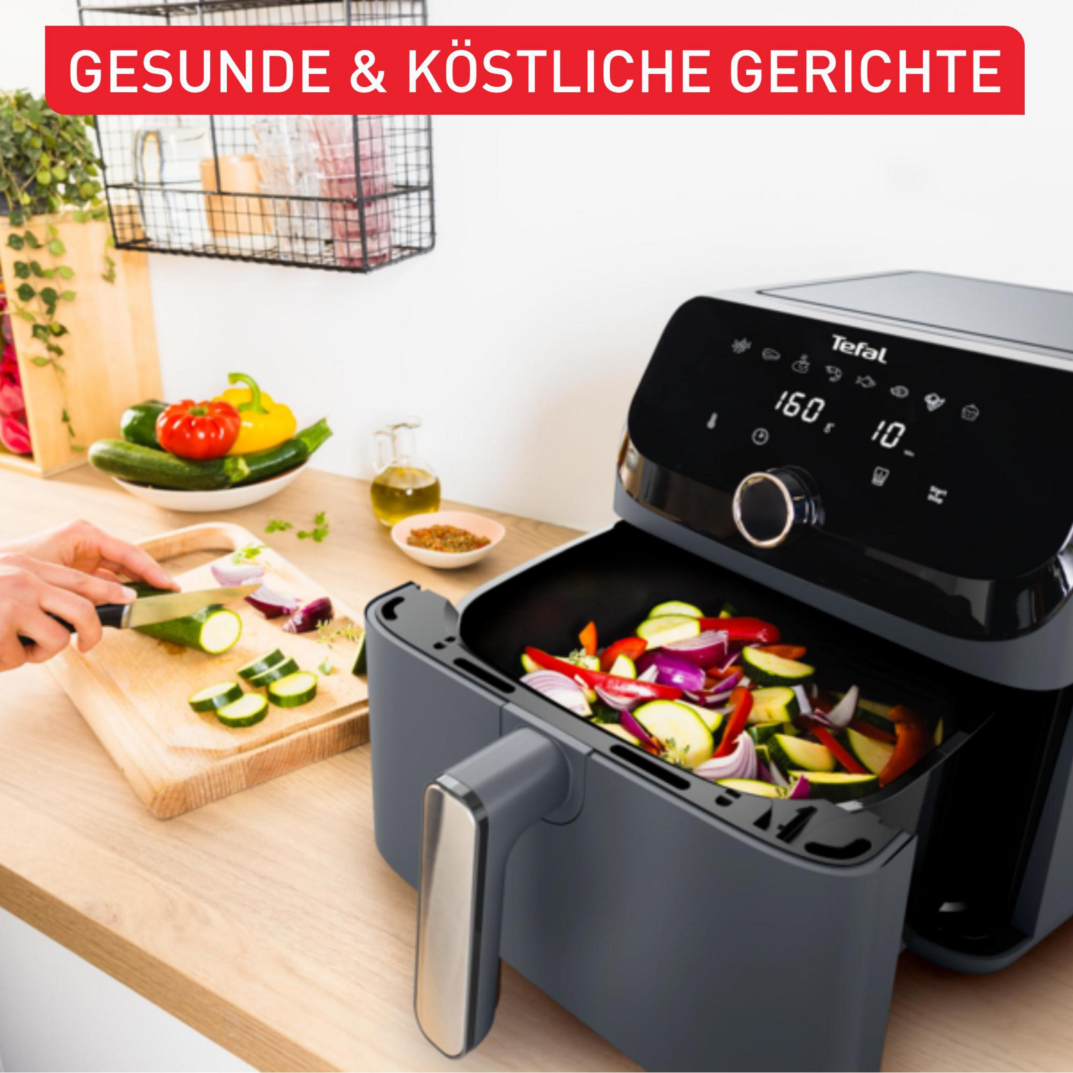 Eine Person schneidet Zucchini auf einem Holzbrett, neben einer Heißluftfritteuse mit Gemüse.