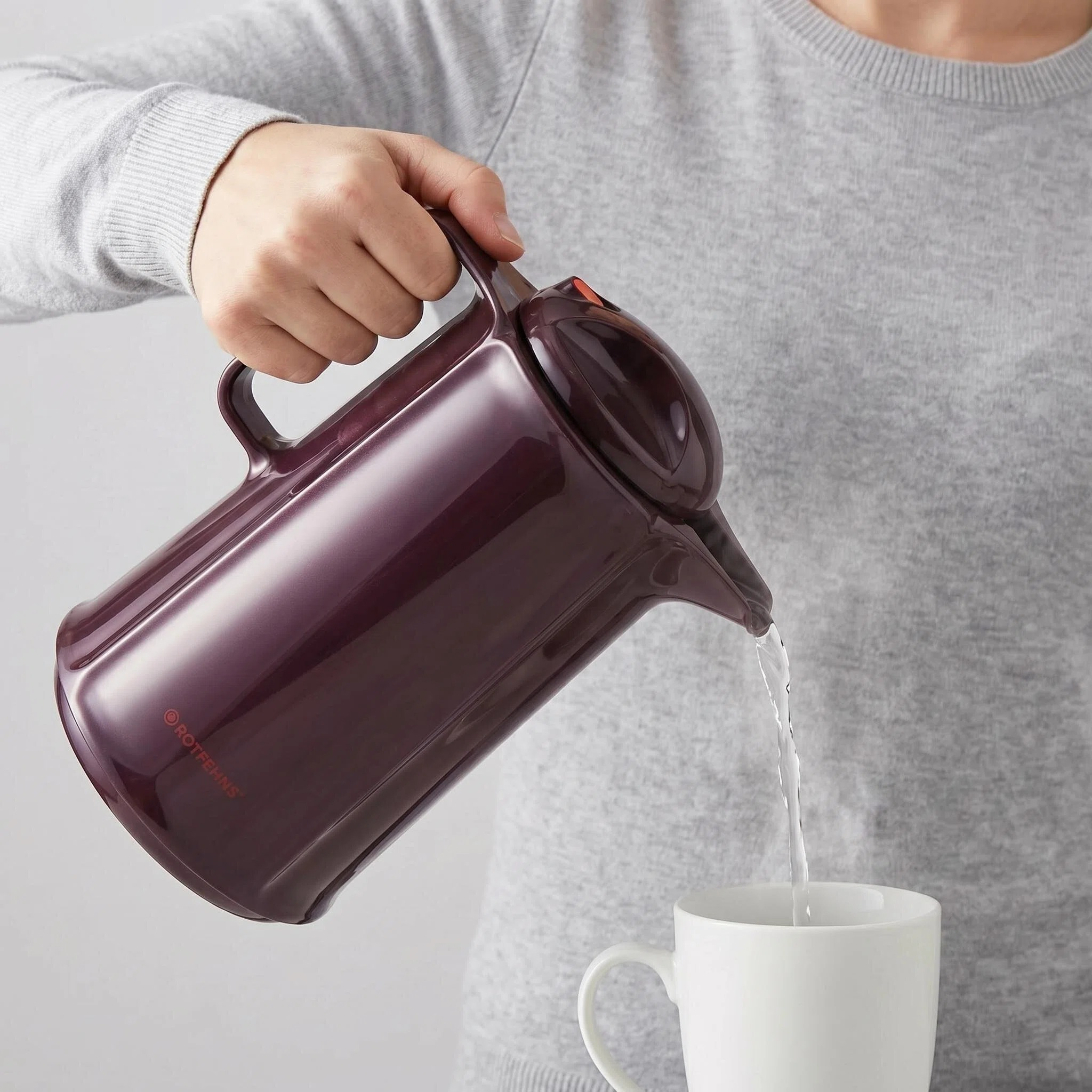 Eine Person gießt mit einer dunkelroten Kanne Wasser in eine weiße Tasse.