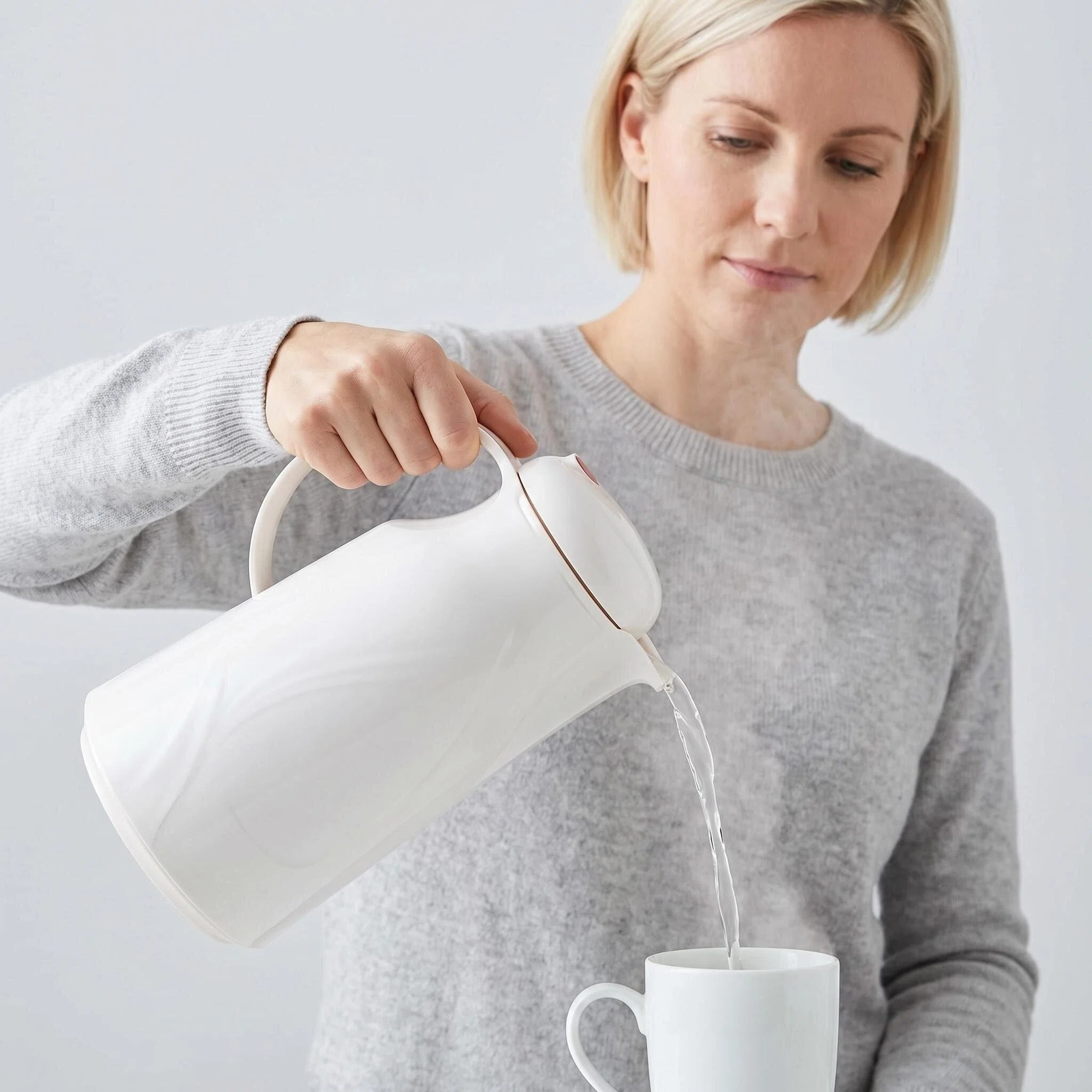 Eine Frau gießt Wasser aus einer weißen Kanne in eine weiße Tasse.
