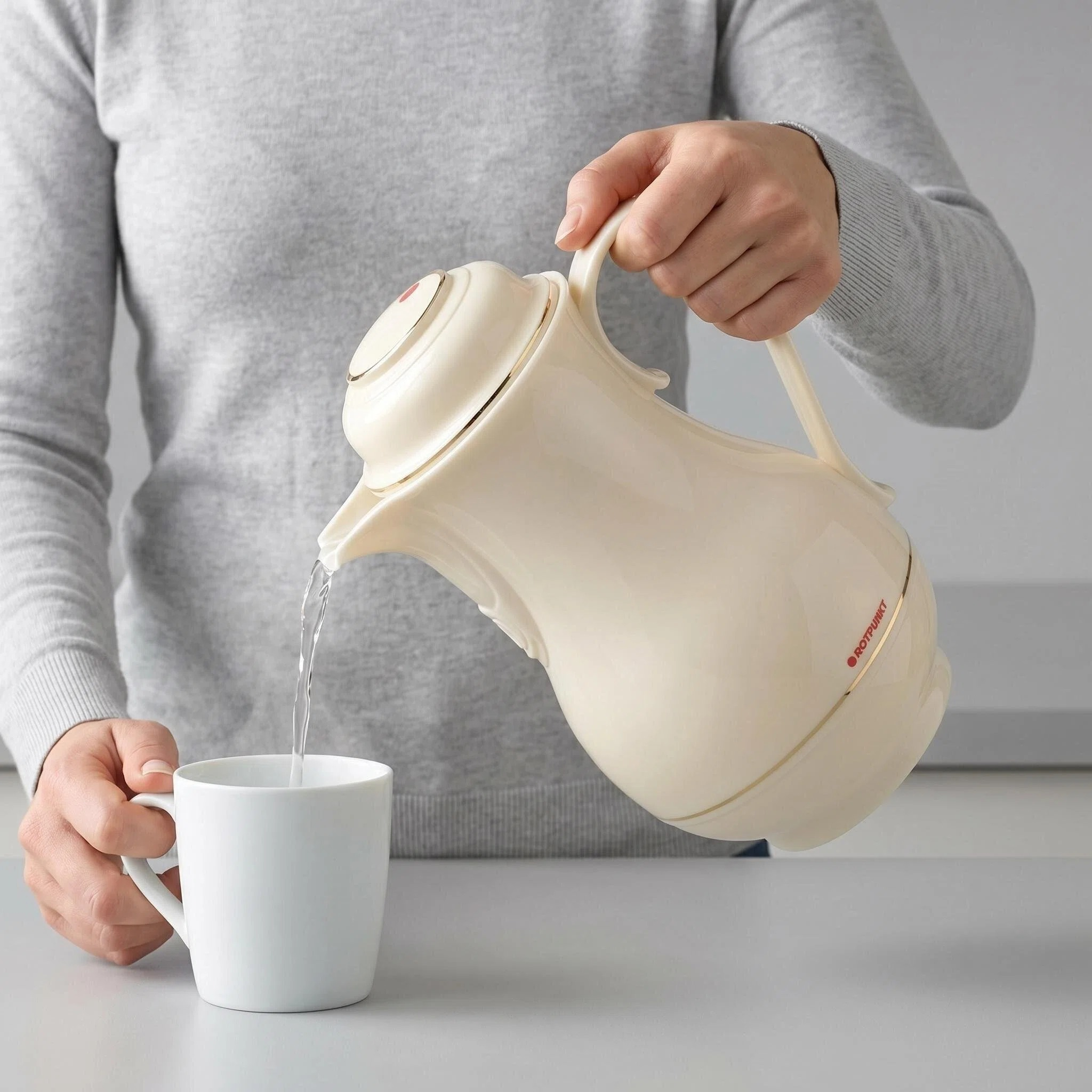 Eine Person gießt Wasser aus einer cremefarbenen Kanne in eine weiße Tasse.