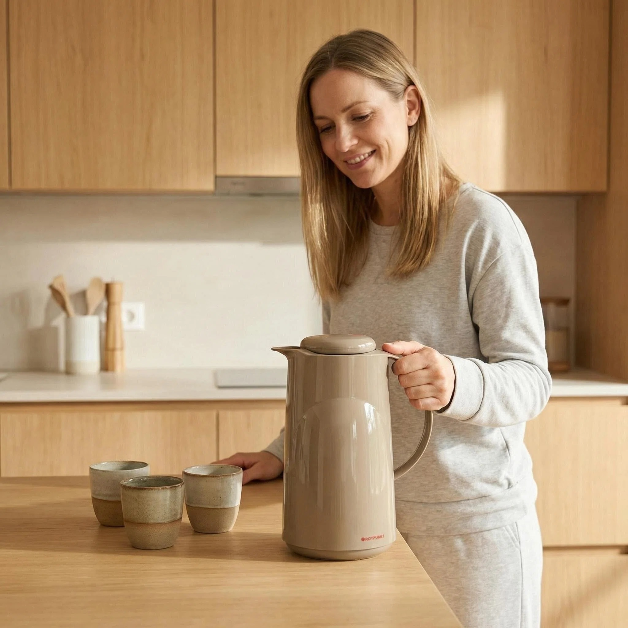 Una donna afferra un thermos beige accanto a tre tazze su un tavolo di legno.