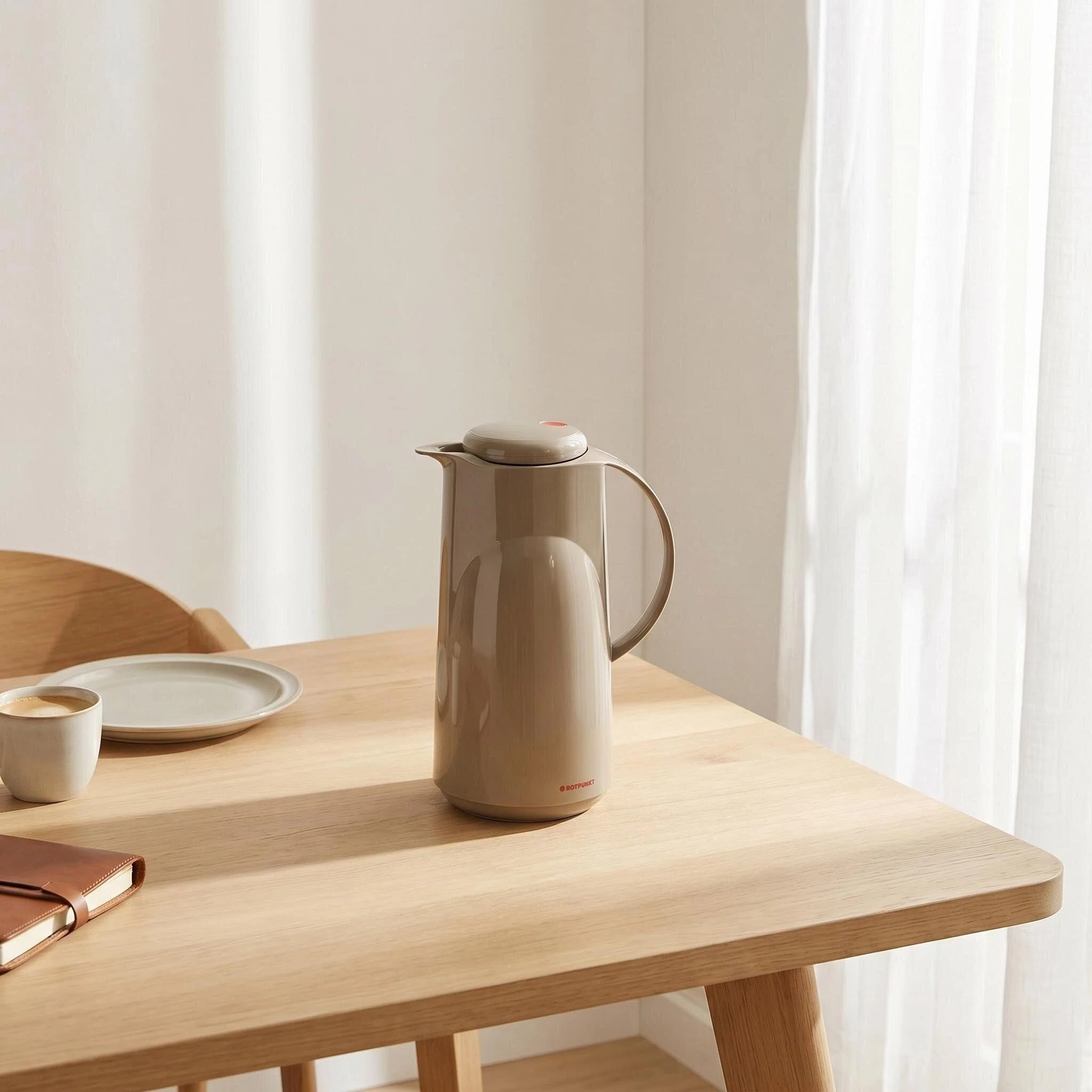 Una brocca termica beige è su un tavolo di legno accanto a una tazza di caffè.