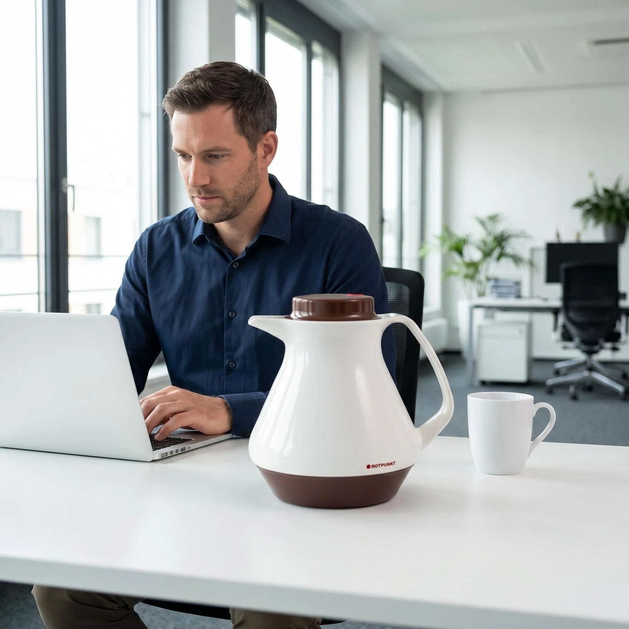 Mann arbeitet am Laptop, neben ihm ein weißer Thermoskanne und eine Tasse.