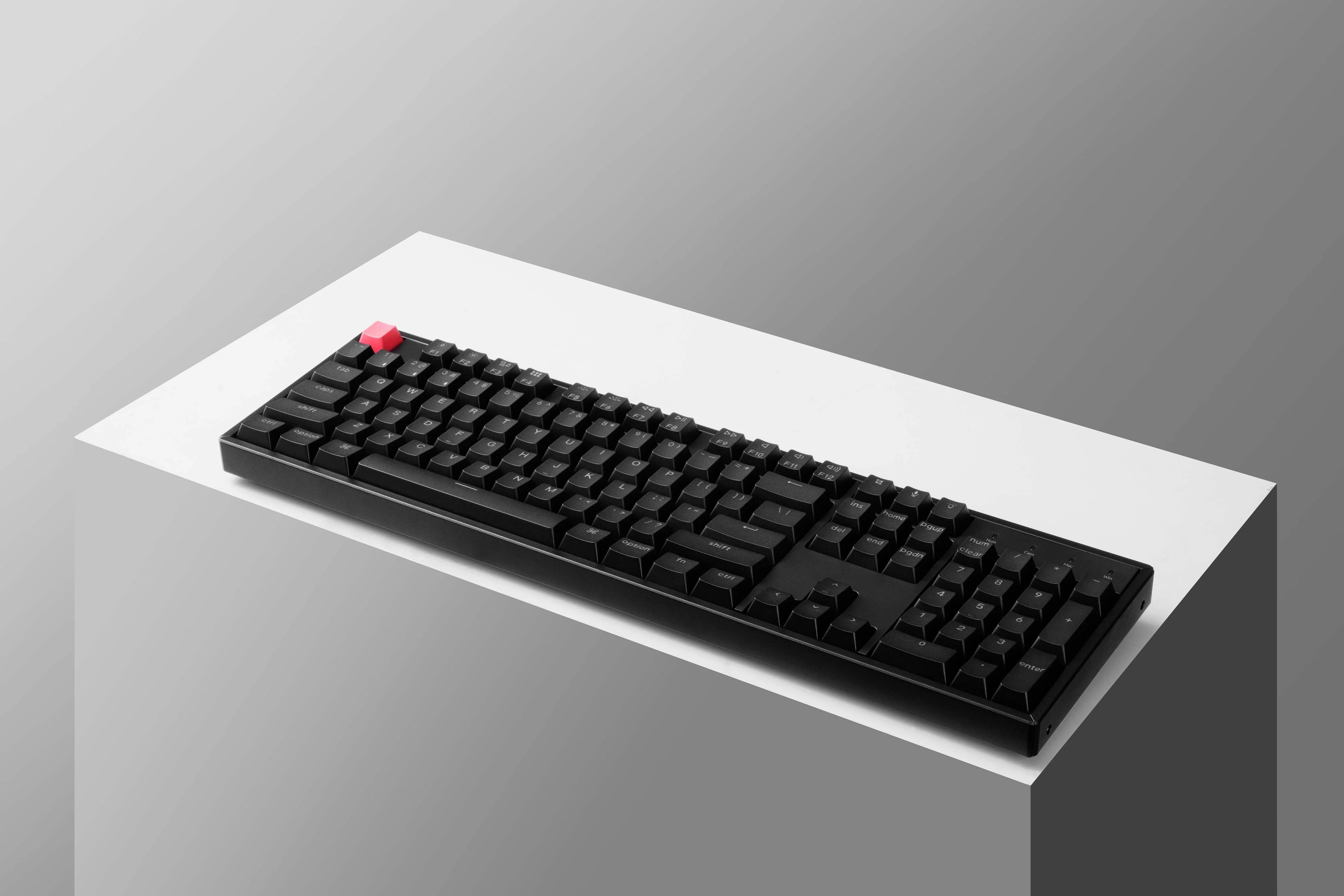 Czarna klawiatura mechaniczna z czerwonym klawiszem na białej powierzchni na szarym tle.