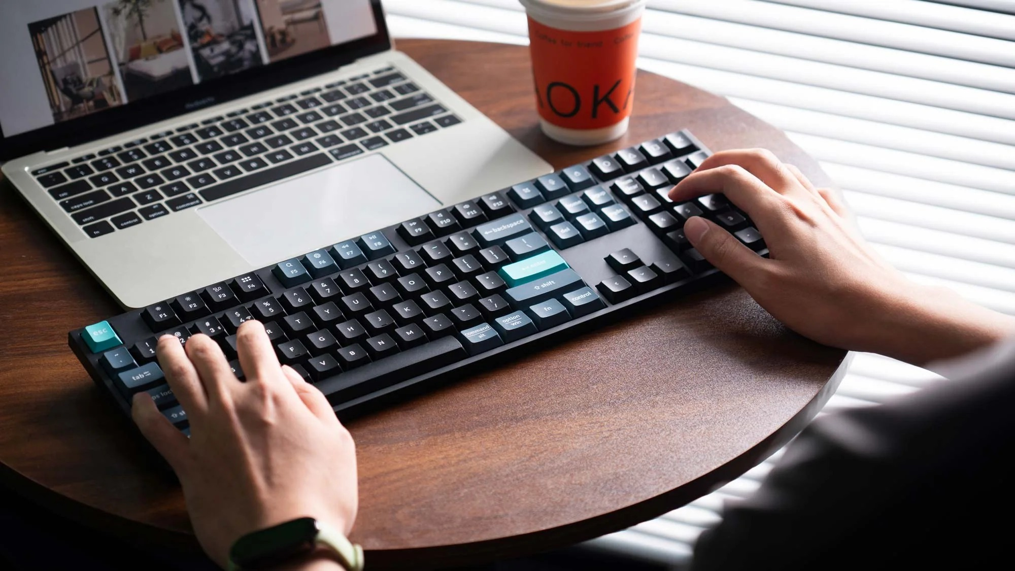 Dłonie piszą na czarnej klawiaturze obok laptopa i pomarańczowego kubka.
