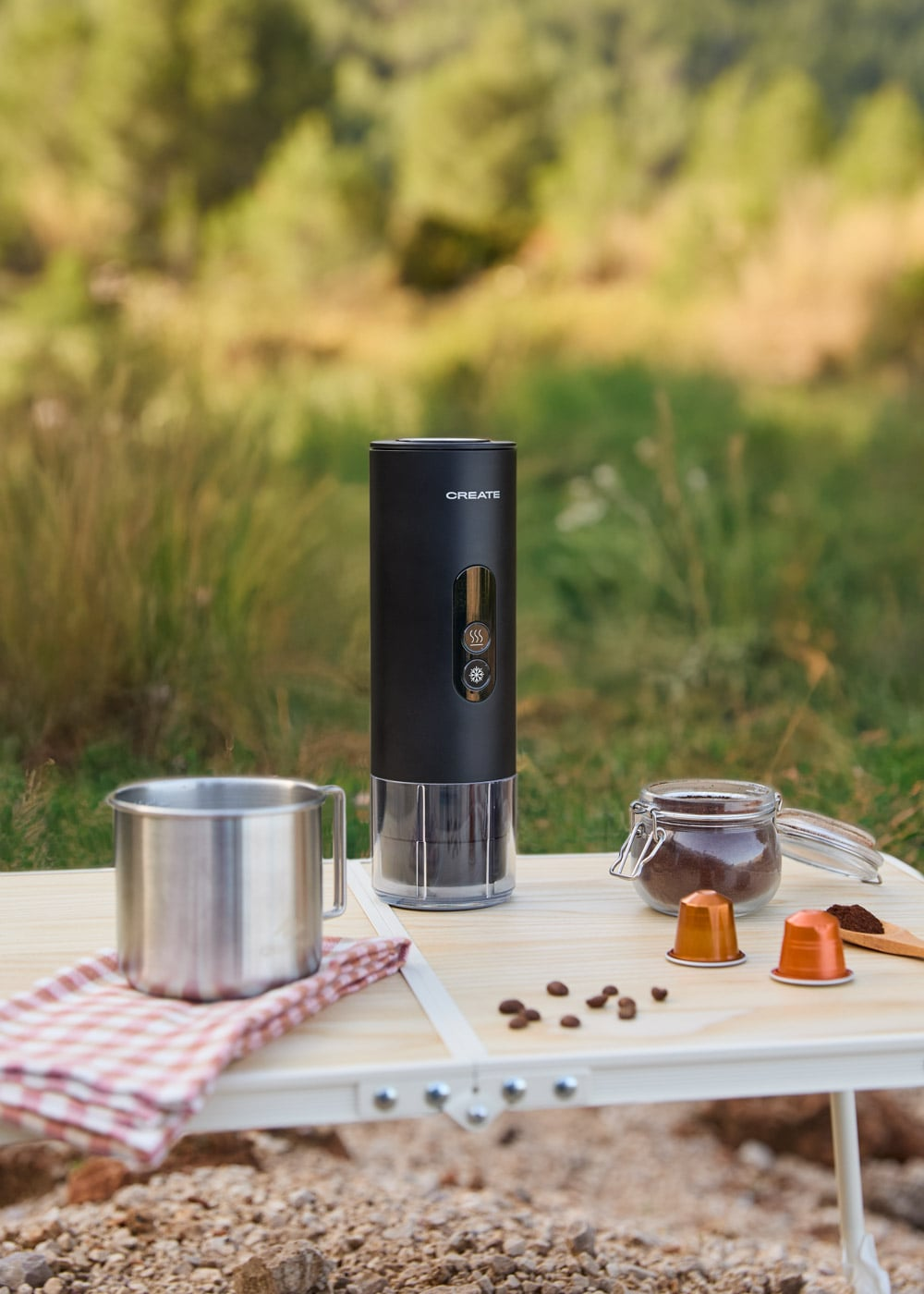 Una cafetera portátil negra está sobre una mesa al aire libre con una taza de metal y posos de café.