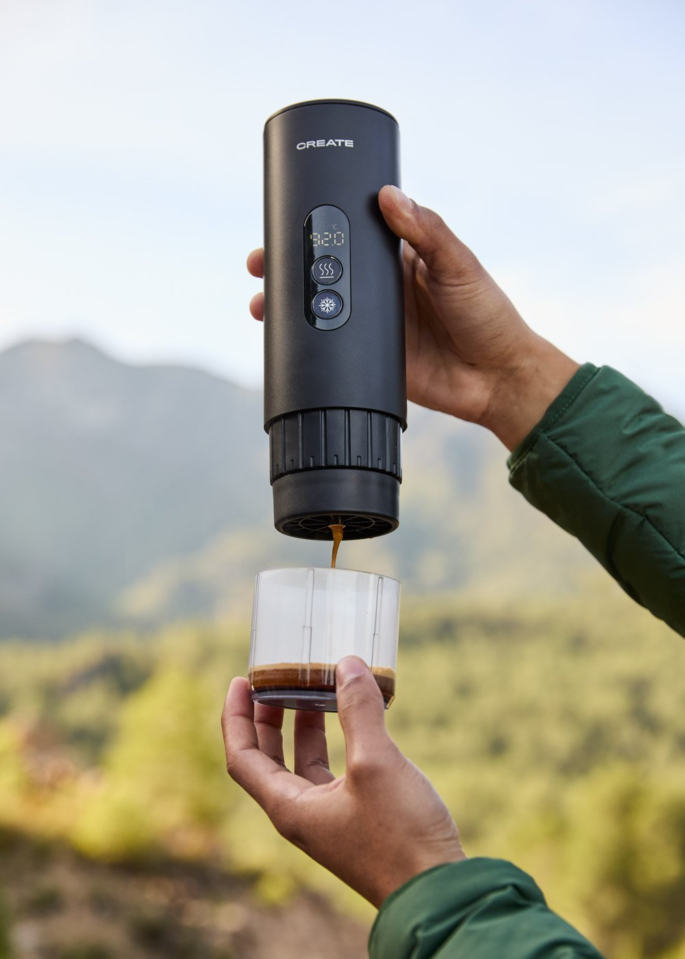 Una mano sostiene una cafetera negra vertiendo café en un vaso. Al aire libre.