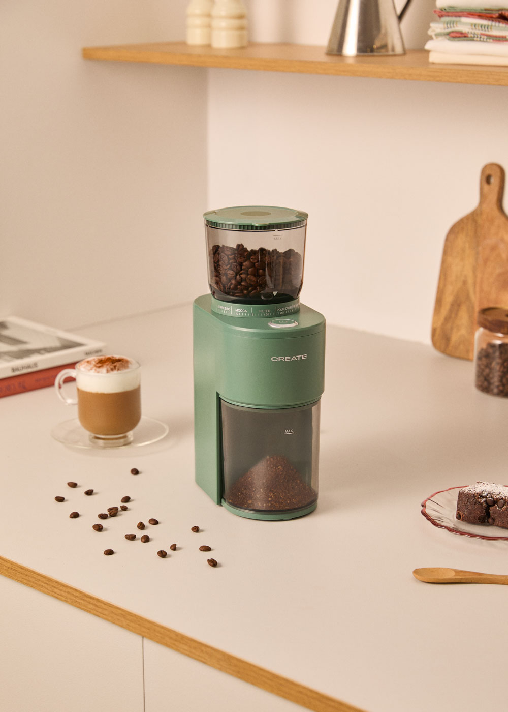 Molinillo de café verde con granos arriba y molido abajo. Cerca hay una taza de latte.