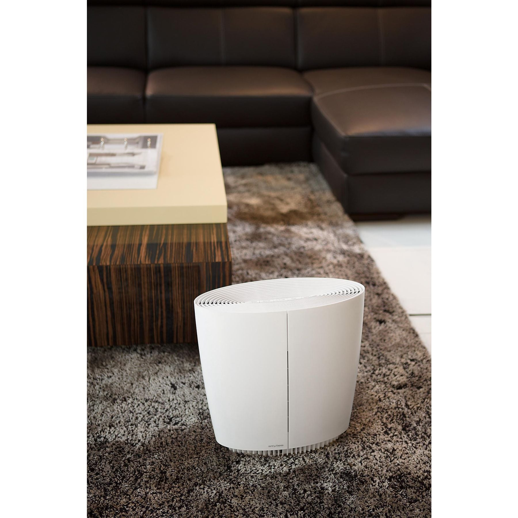 Ein weißer Luftreiniger steht auf einem flauschigen Teppich neben einem Couchtisch und Sofa.