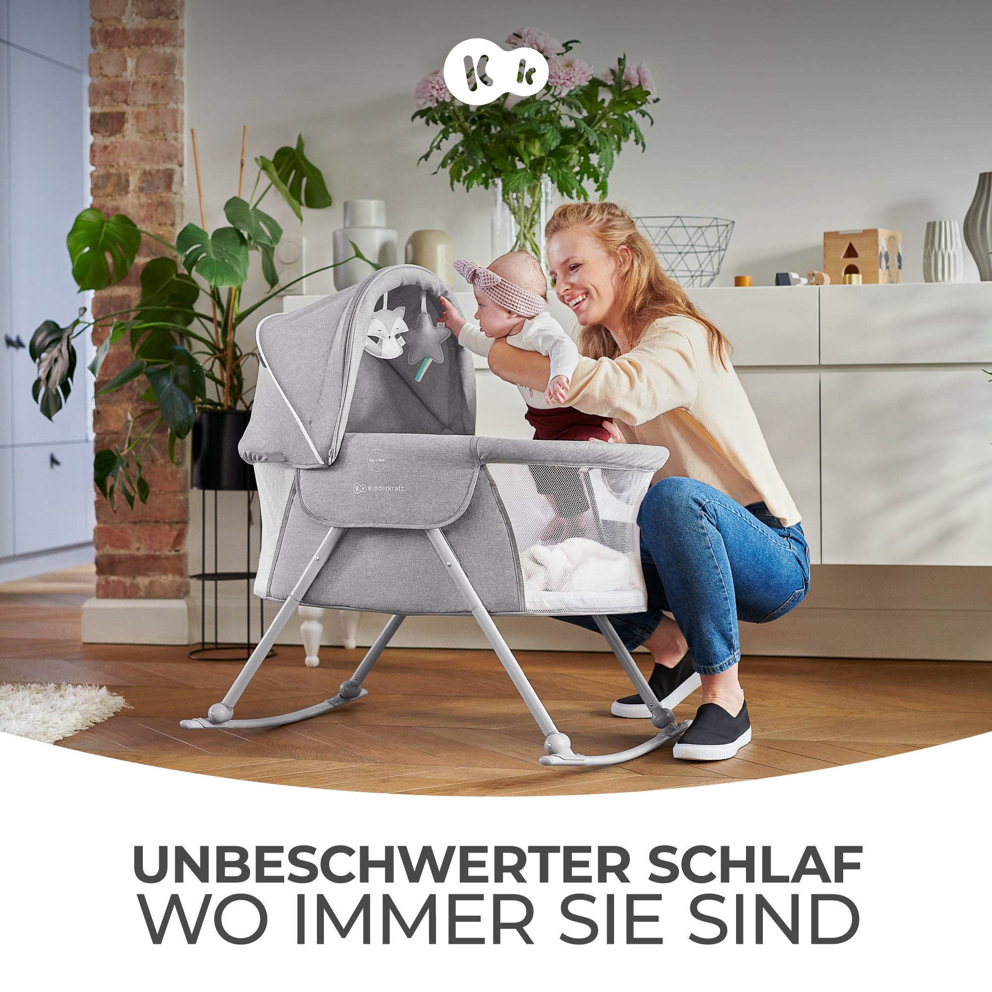 Eine Frau hockt neben einem grauen Babybett mit einem Spielzeug mobile, hält ein Baby.