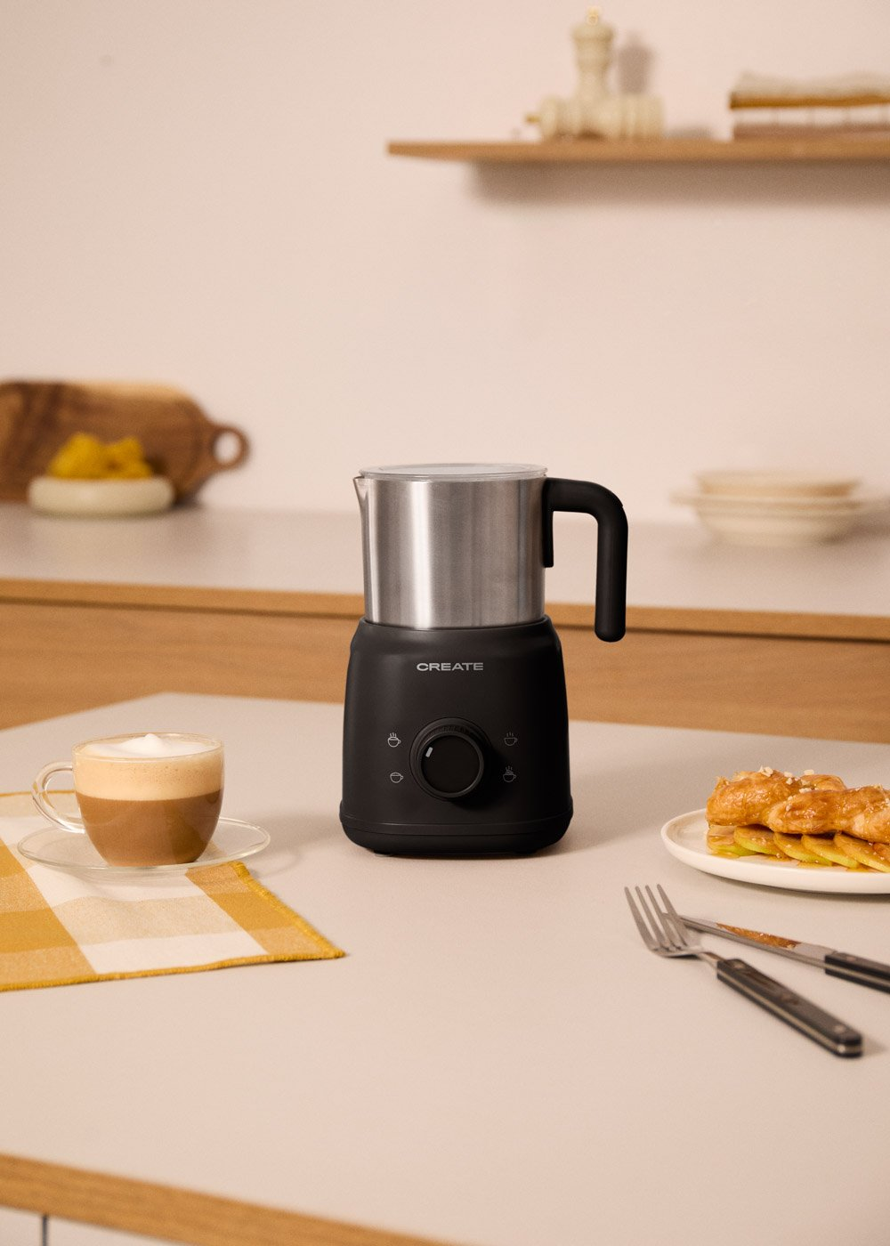 Un espumador de leche negro junto a una taza de café y un plato de pasteles.