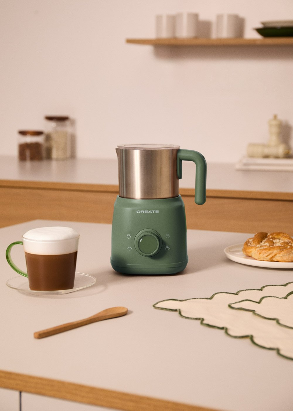 Un espumador de leche verde con jarra de acero inoxidable junto a una taza de café y un pastel.