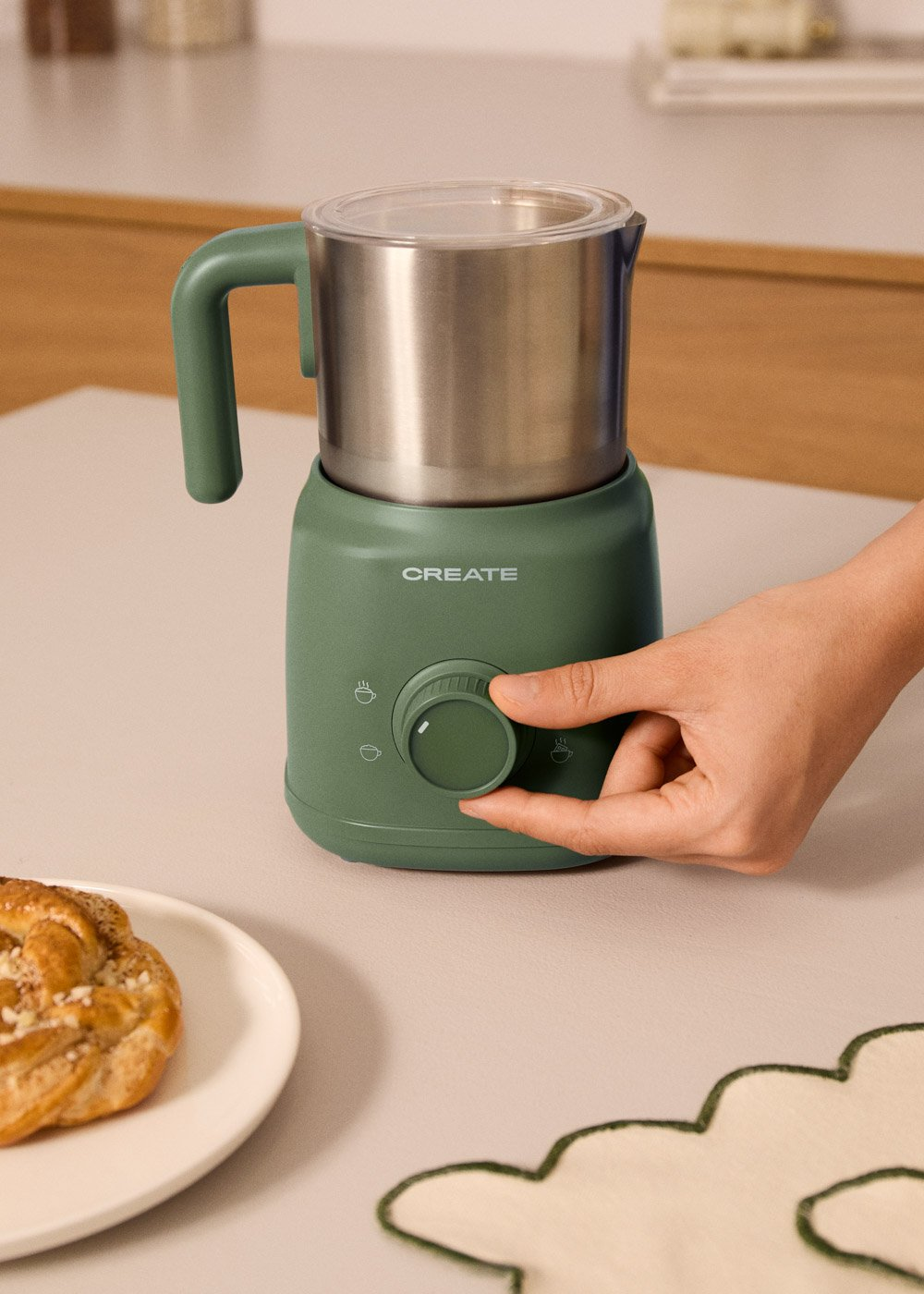 Un espumador de leche verde con jarra de acero inoxidable. Una mano ajusta el dial.