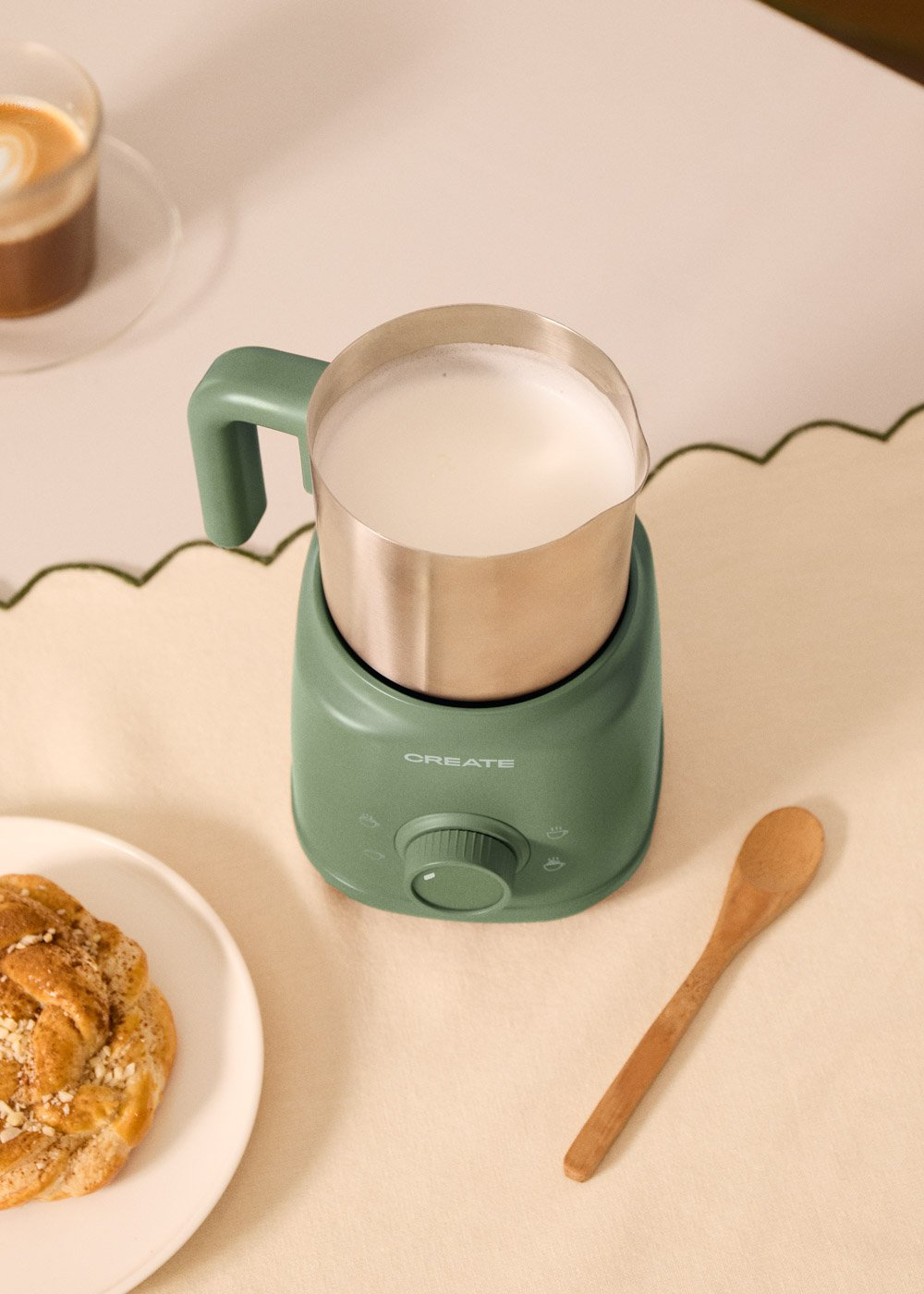 Un espumador de leche verde con leche espumada, al lado un pastel y una cuchara de madera.