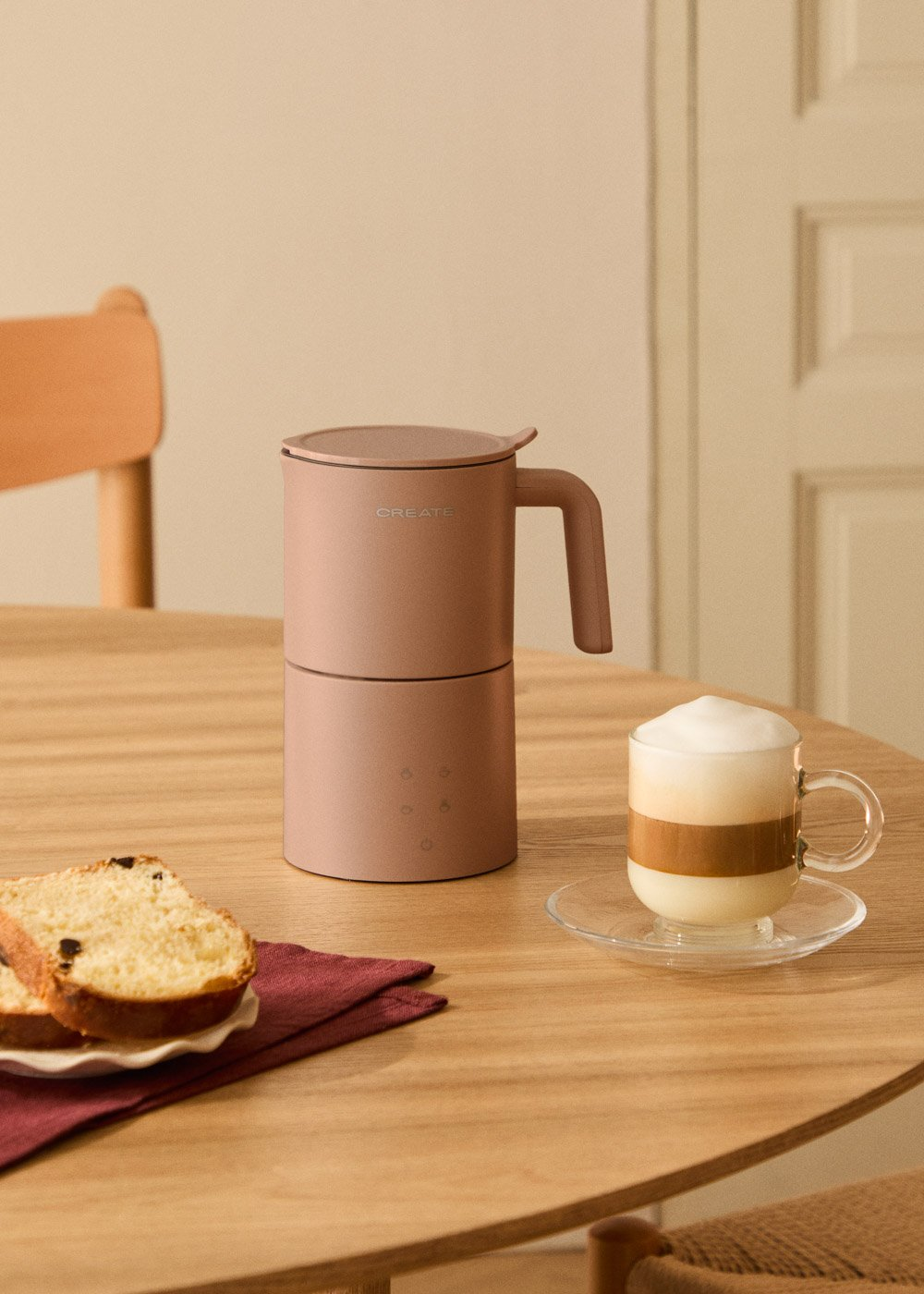 Una cafetera rosa está sobre una mesa de madera con rebanadas de pan y un café con leche.