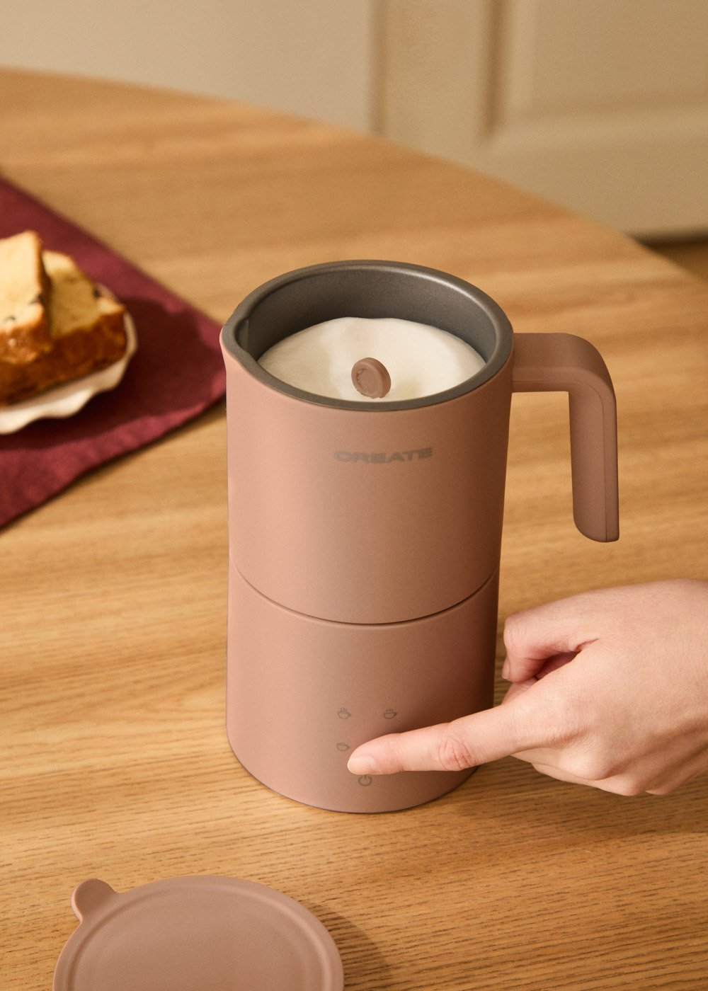 Una mano presiona un botón en un espumador de leche rosa con espuma dentro. Hay trozos de pastel cerca.