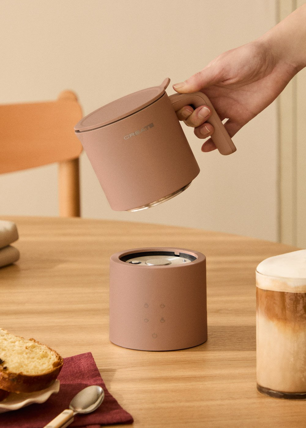 Una mano sostiene una taza rosa sobre una base a juego en una mesa de madera con pan y café.