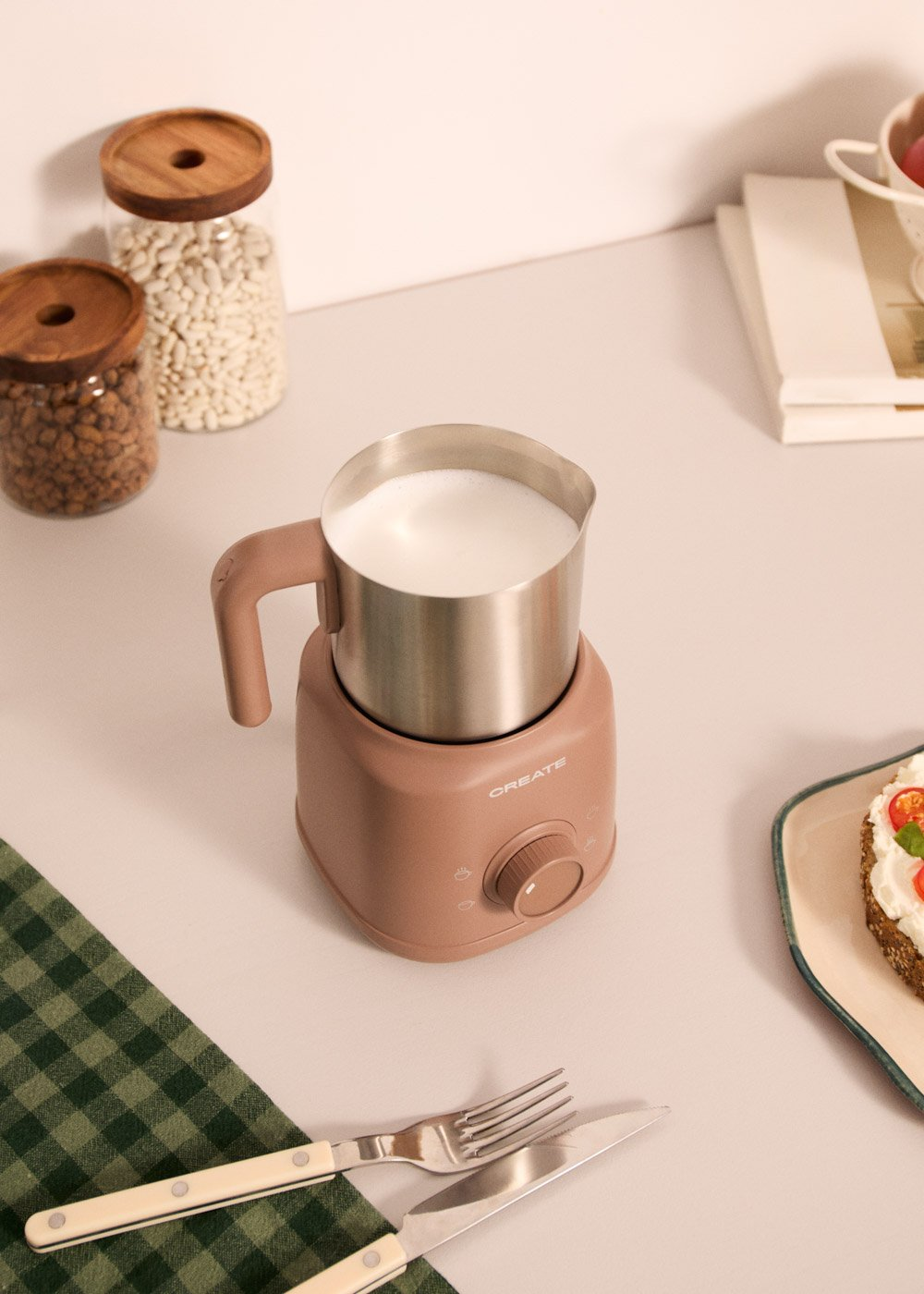 Espumador de leche con leche, dos tarros de frijoles, cubiertos y tostada en mesa.