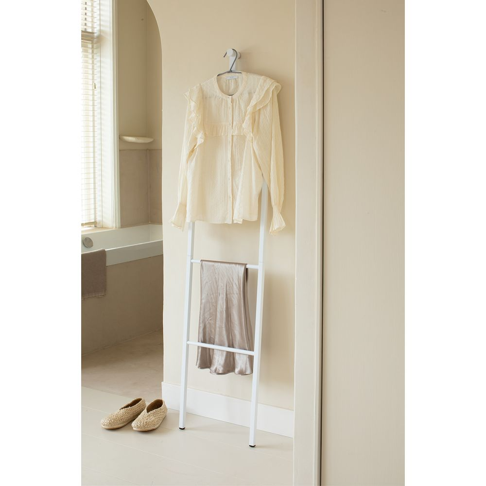 Eine cremefarbene Bluse und ein beiger Rock hängen auf einem weißen Leiternständer. Zwei Strohschuhe stehen auf dem Boden.