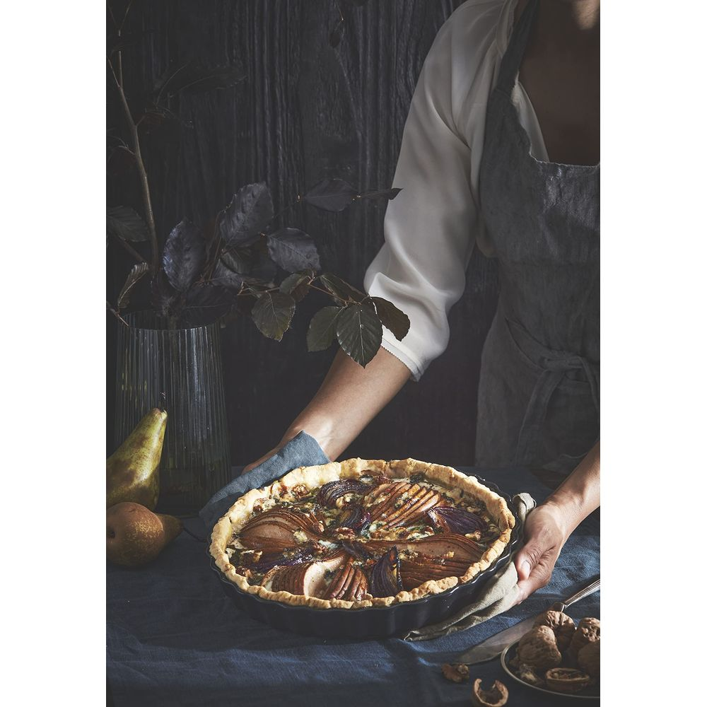 Una persona con grembiule tiene una crostata di pere con noci e rosmarino su una tovaglia blu.