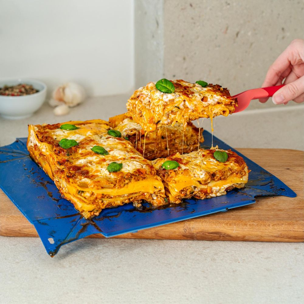 Eine Portion Lasagne wird mit einem roten Löffel aus einer größeren Lasagne gehoben.