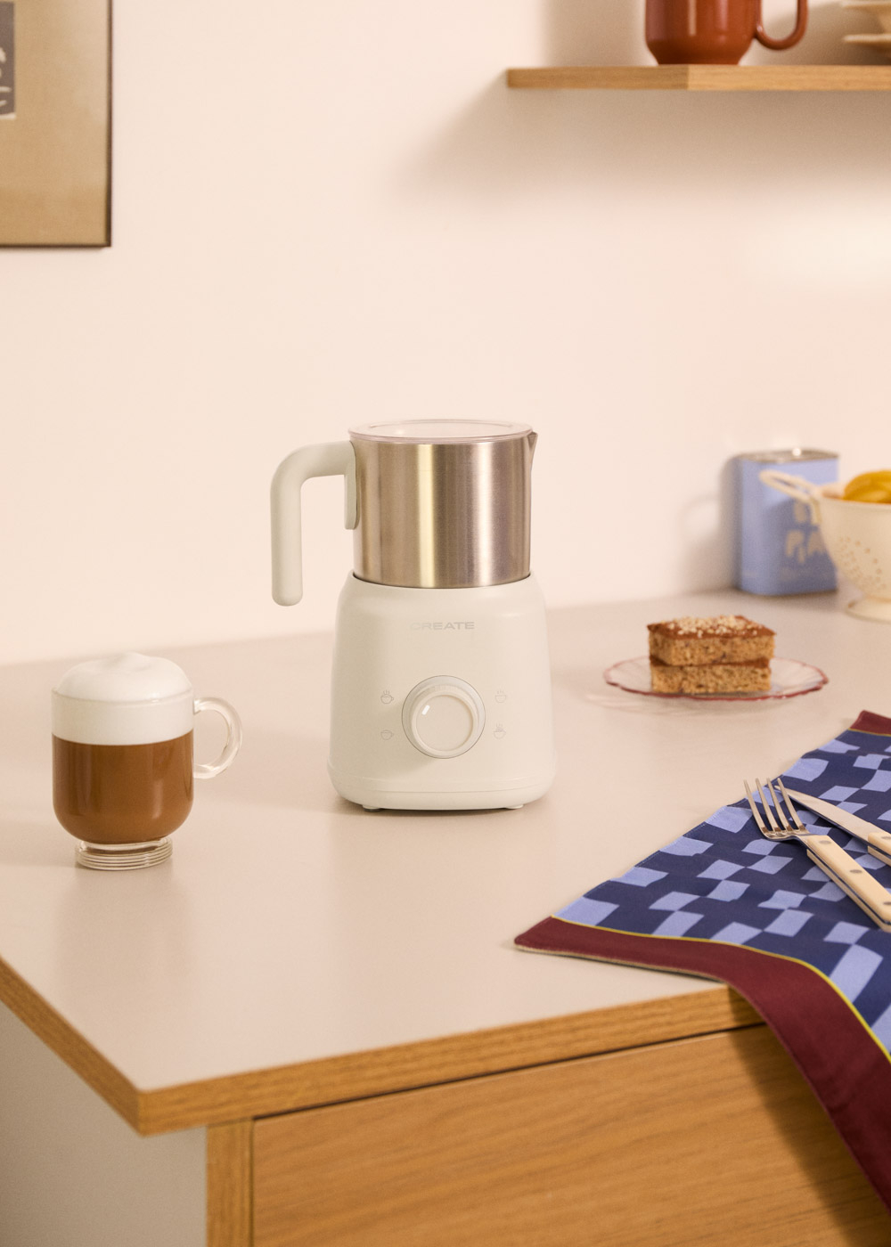 Un espumador de leche blanco con jarra de acero inoxidable junto a un vaso de café.