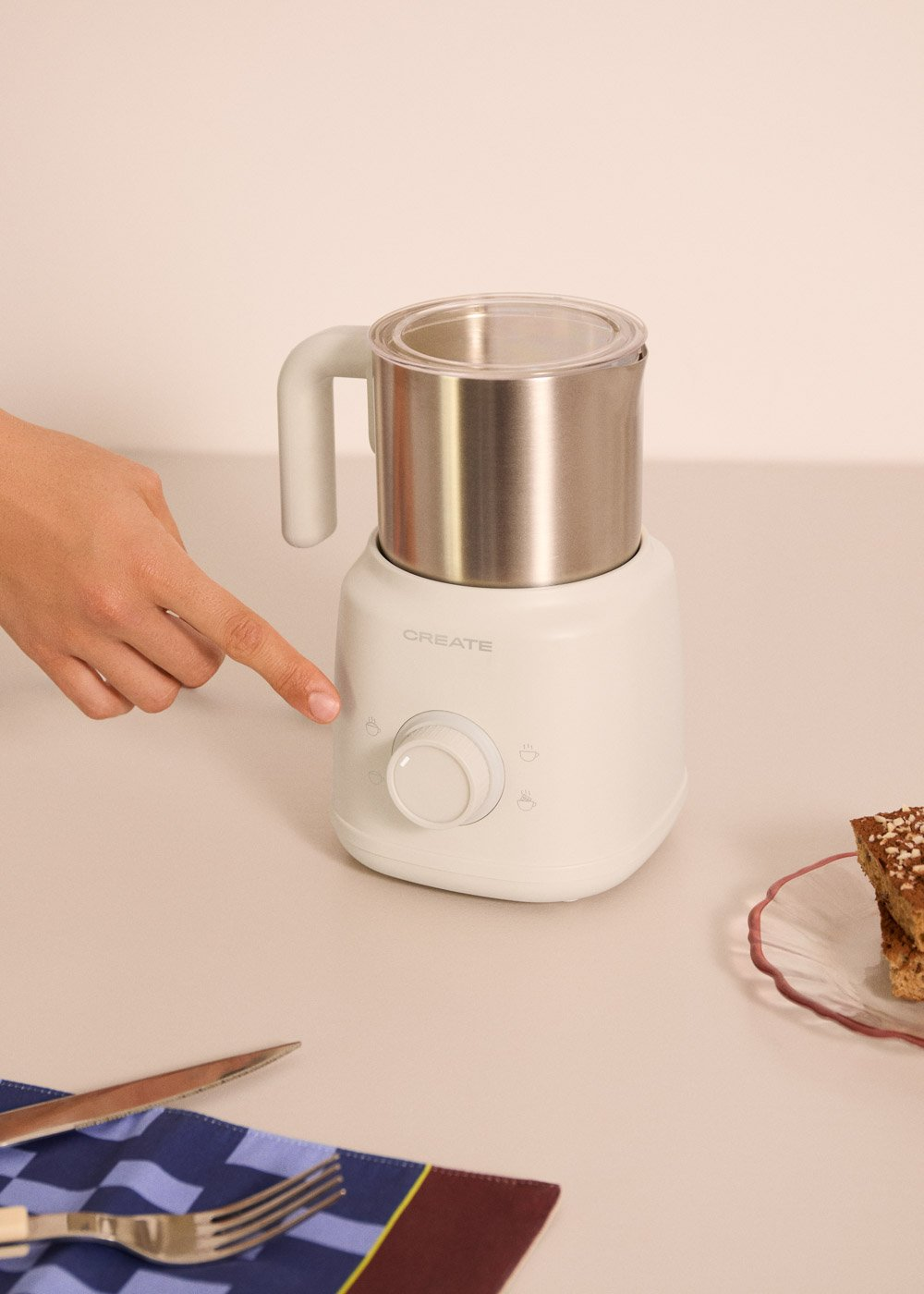 Un electrodoméstico de cocina blanco con jarra metálica y tapa de cristal. Una mano pulsa un botón.