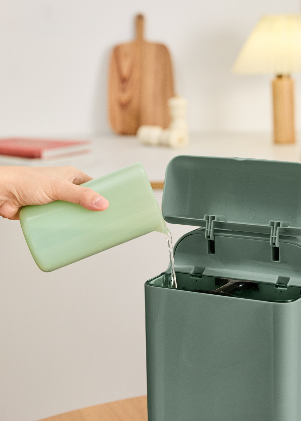 Una mano vierte agua de un vaso verde en un cubo de basura verde abierto.