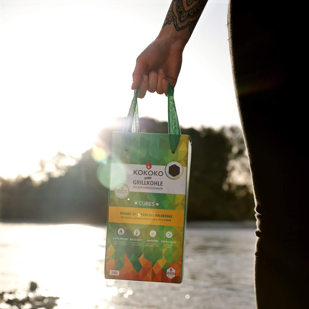 Persona tiene sacchetto carbone KOKOKO con manici verdi, acqua soleggiata sullo sfondo.