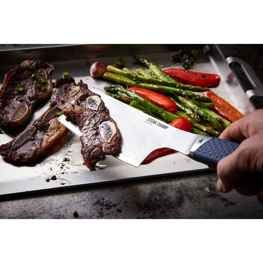 Una mano usa una spatola Steak Champ per girare una bistecca con l'osso accanto ad asparagi e peperoni rossi grigliati.