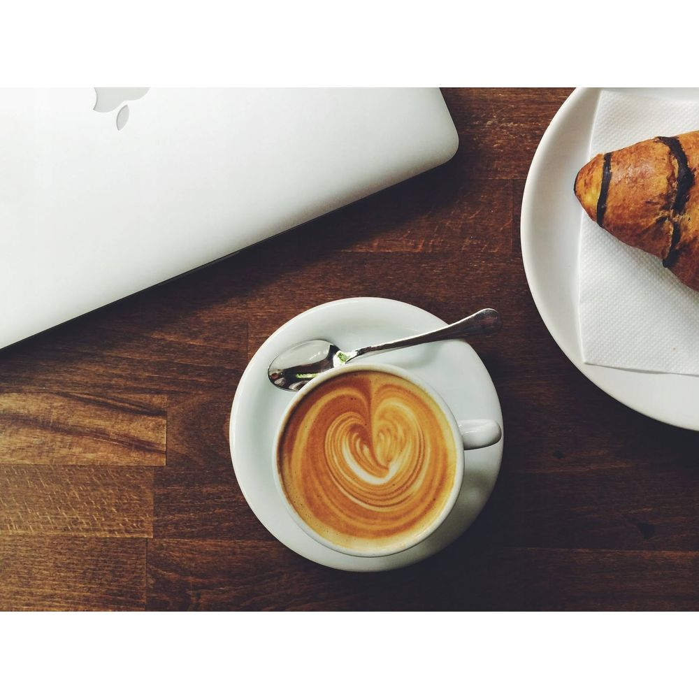 Un laptop, una tazza di caffè latte art e un croissant su un tavolo di legno.