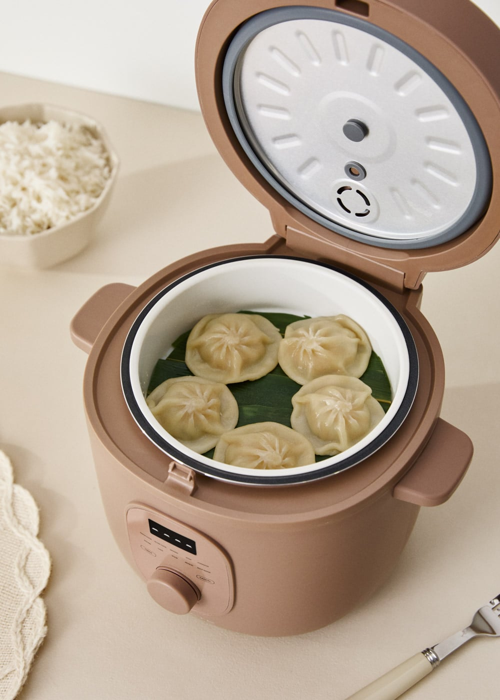 Vaporera con cinco empanadillas al vapor sobre hojas verdes, junto a un bol de arroz.