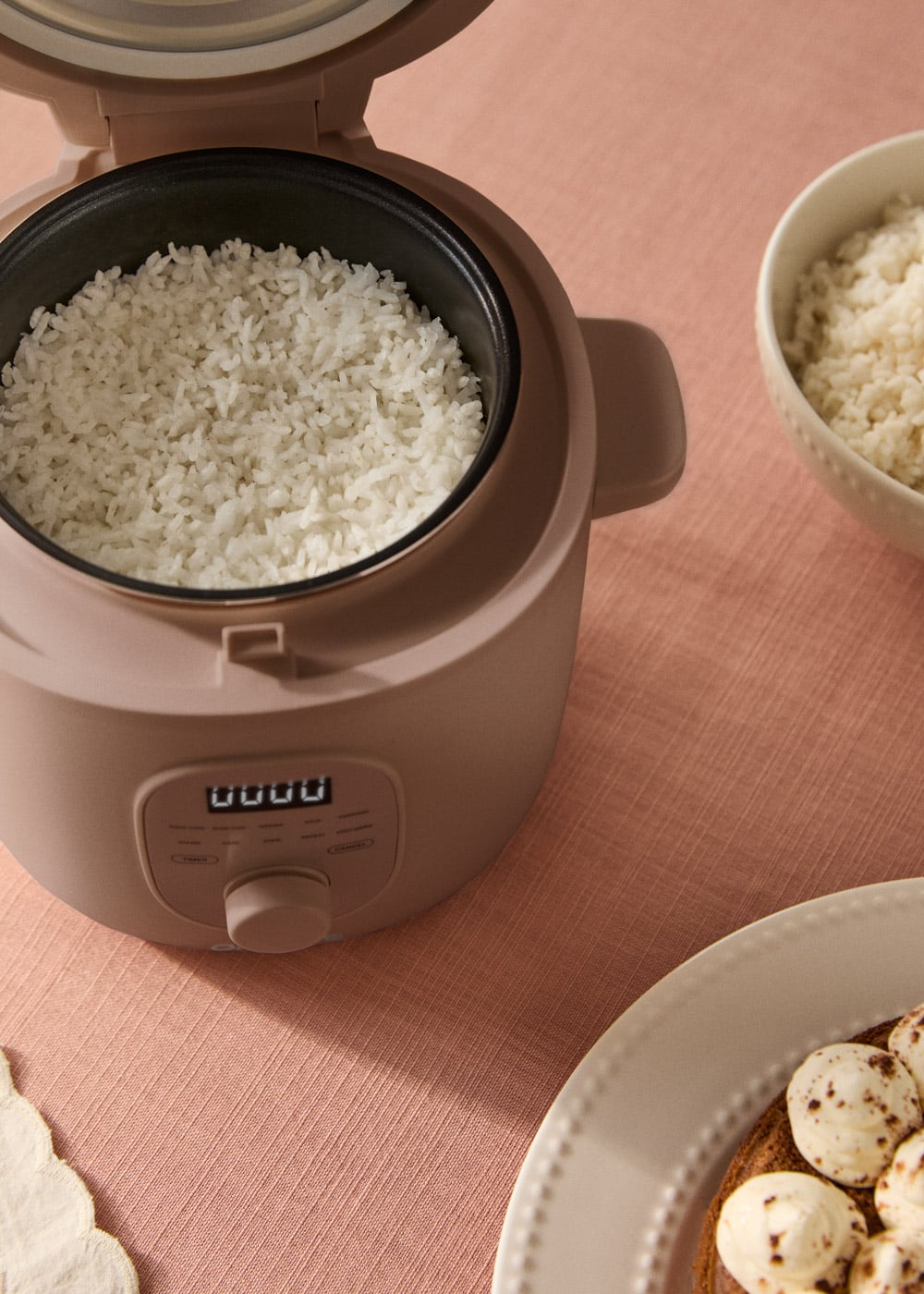 Una arrocera abierta llena de arroz cocido, junto a un bol de arroz sobre un mantel rosa.