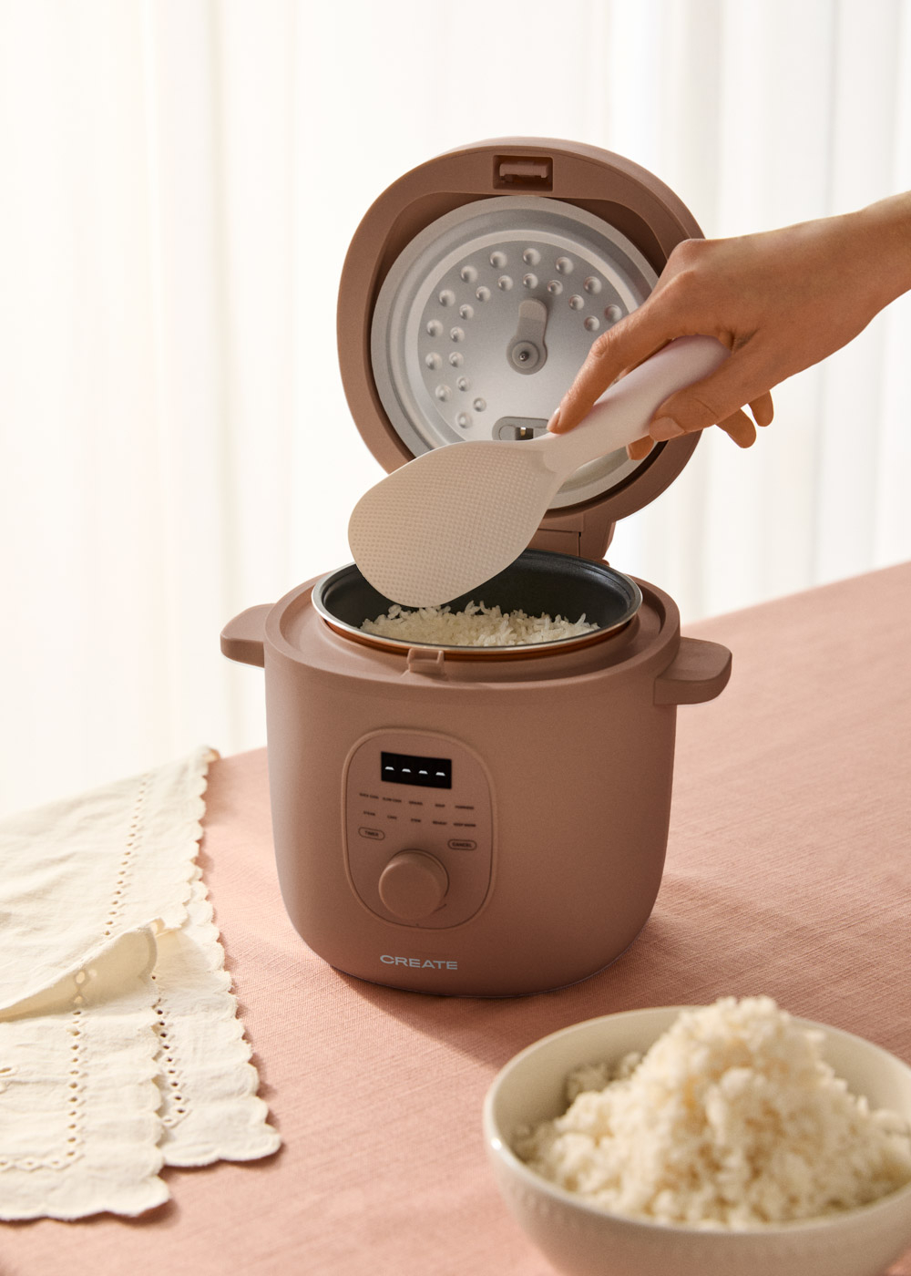 Una mano usa una espátula de arroz para sacar arroz de una arrocera. Hay un bol de arroz cerca.