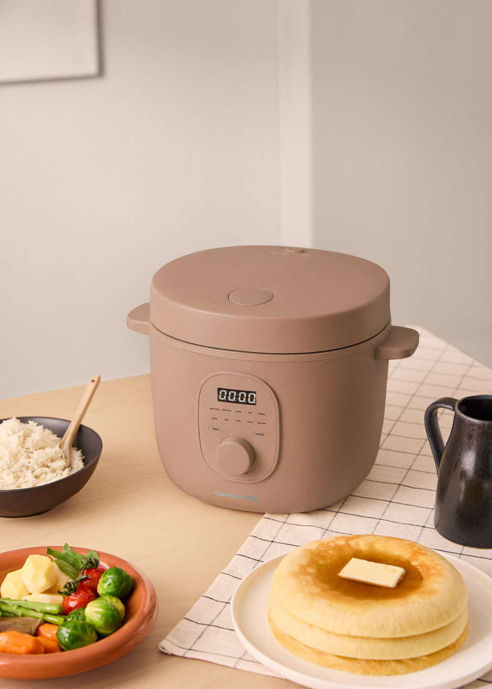 Una multicocina beige está sobre una mesa con arroz, verduras y tortitas.
