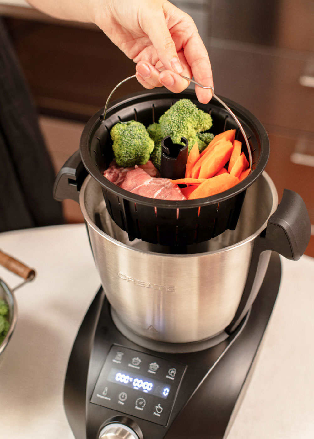 La mano coloca una cesta con carne, brócoli y zanahorias en un procesador de alimentos.