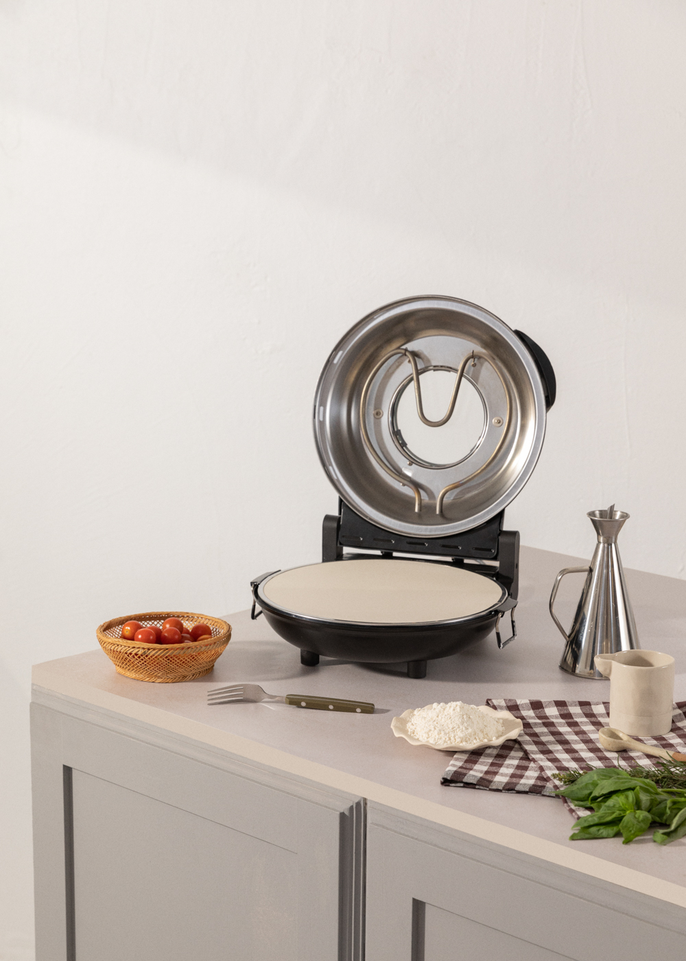 Un horno para pizza con la tapa abierta, junto a un cuenco de tomates cherry y harina.