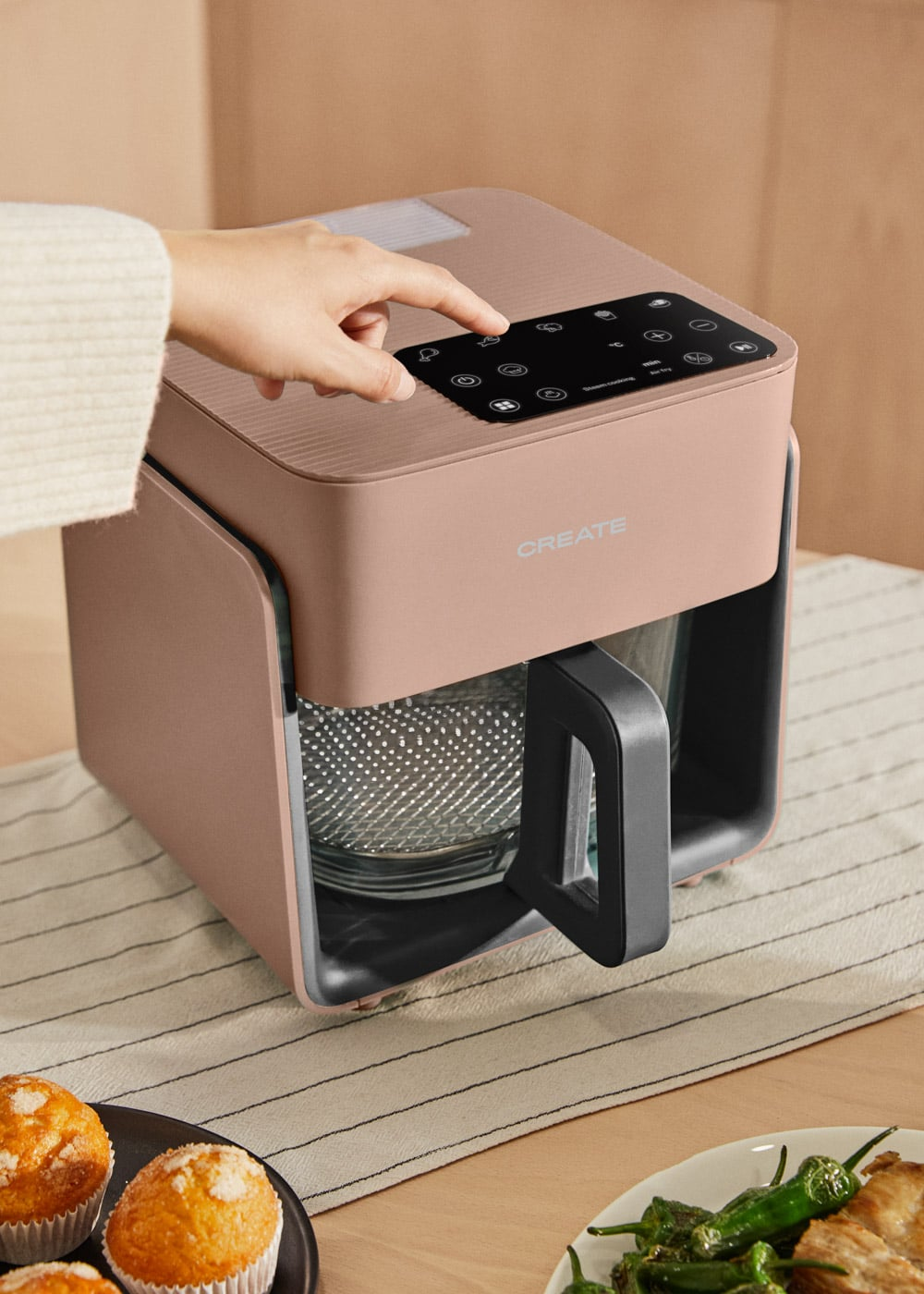 Una freidora de aire oro rosa con pantalla digital es operada por un dedo. Hay muffins en un plato.